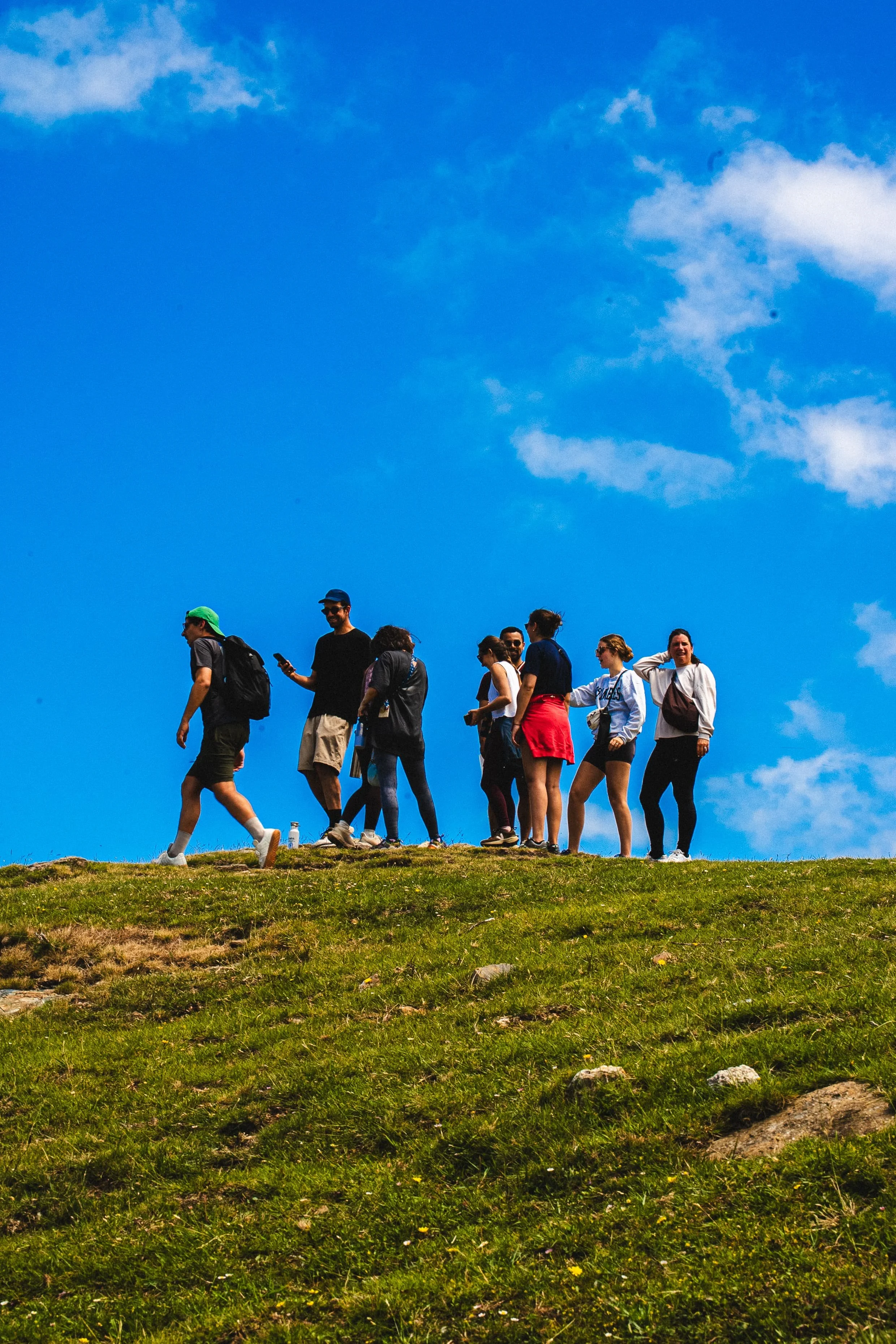 Groupe de personnes marchant sur une colline herbeuse sous un ciel bleu avec quelques nuages.