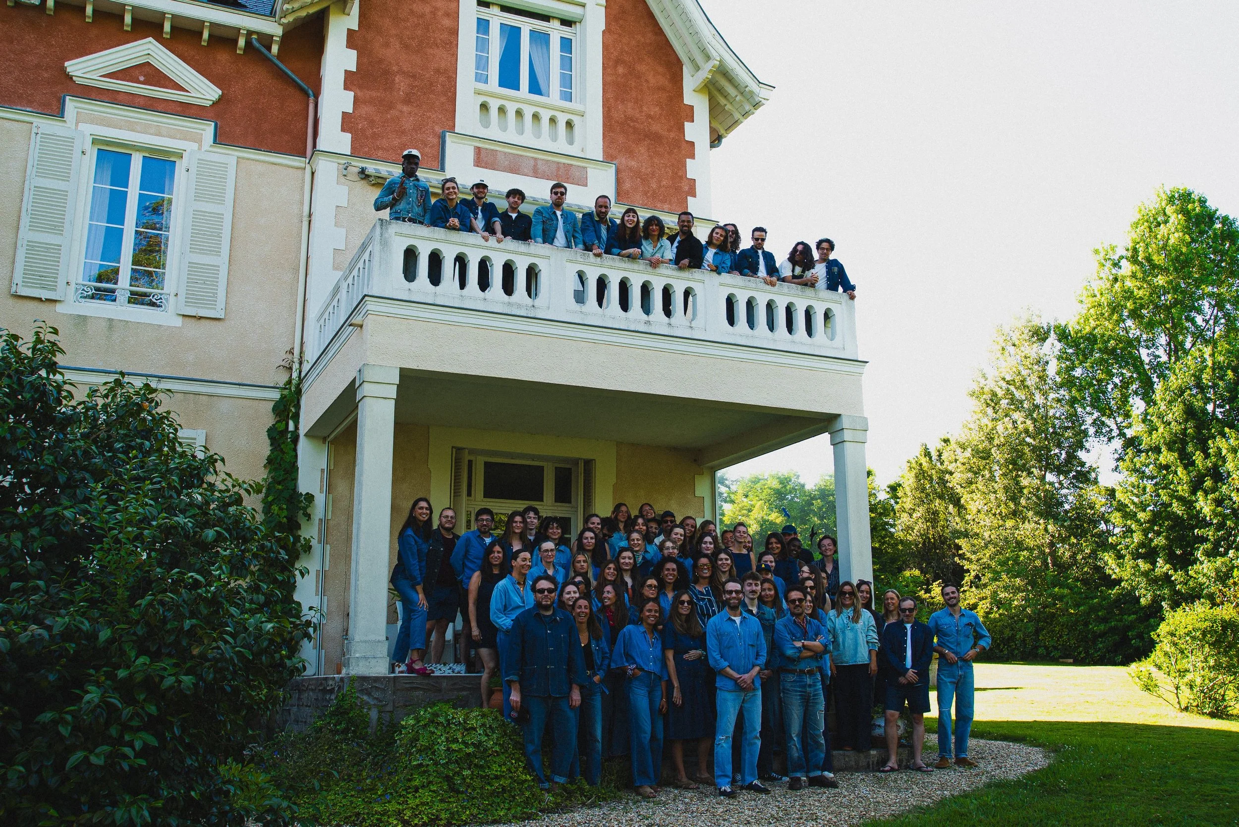 Groupe de personnes posant devant une grande maison avec un balcon. Il y a deux niveaux de personnes, avec des adultes de différentes origines. Tous portent des vêtements bleus ou foncés et sourient, sous un ciel clair avec des arbres verts en arrièr