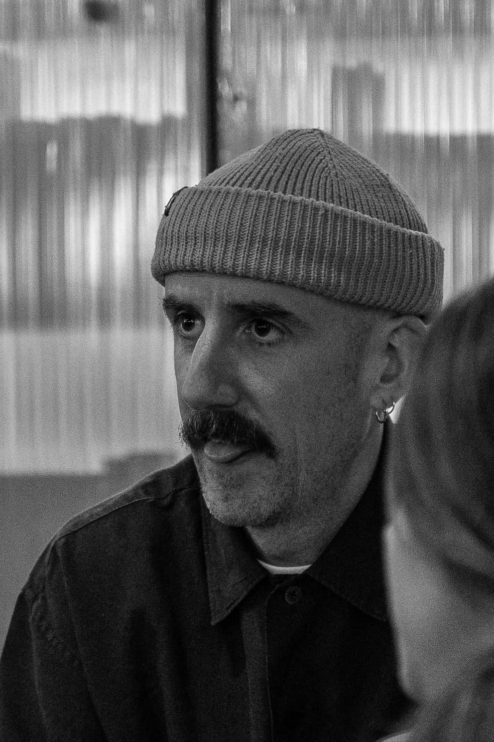 Un homme portant un bonnet en tricot, avec une moustache et une barbe, est en train de parler. L'image est en noir et blanc.