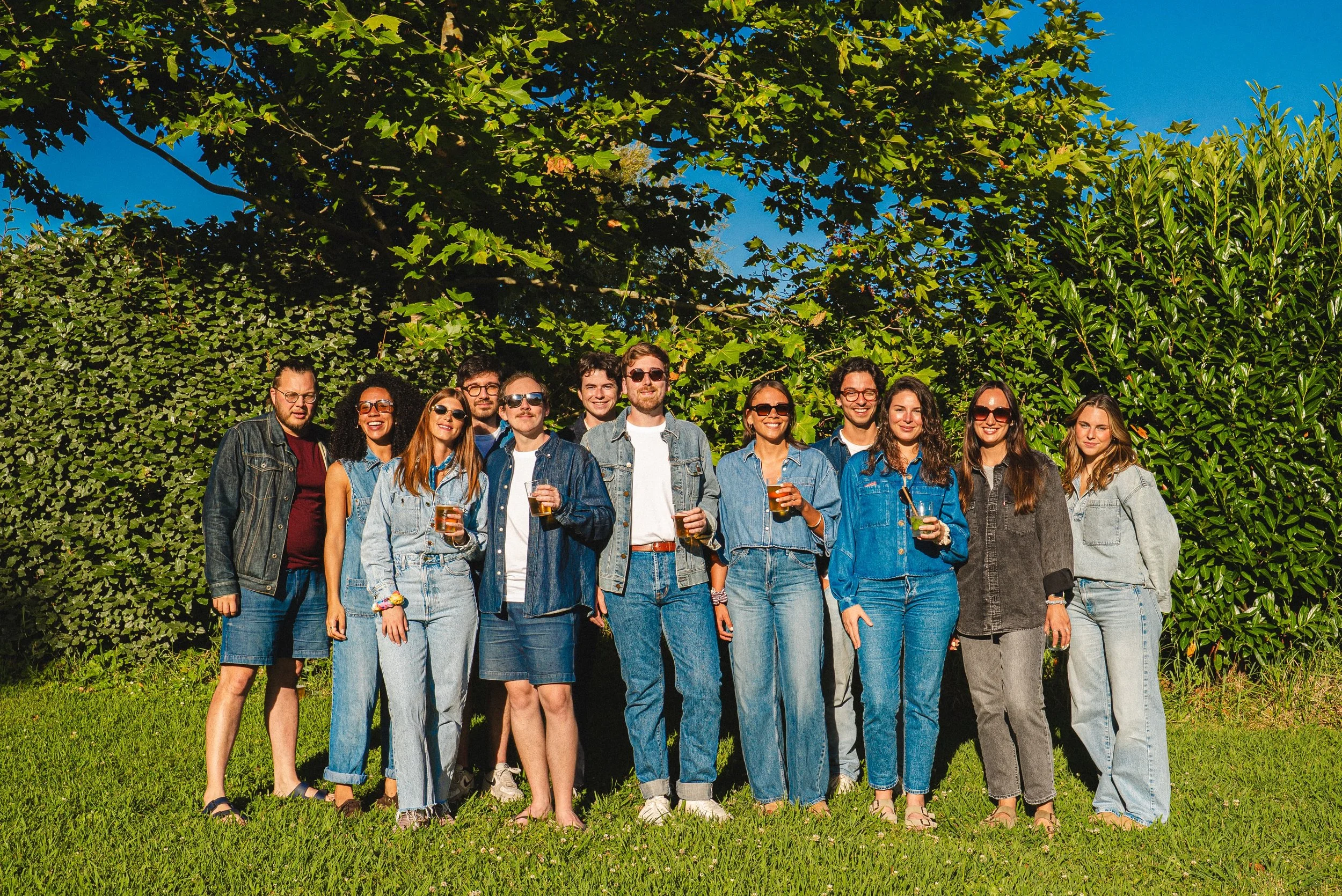 Un groupe de personnes posant dehors devant des buissons et un arbre, tenant des verres de boisson, souriants, en journée ensoleillée.