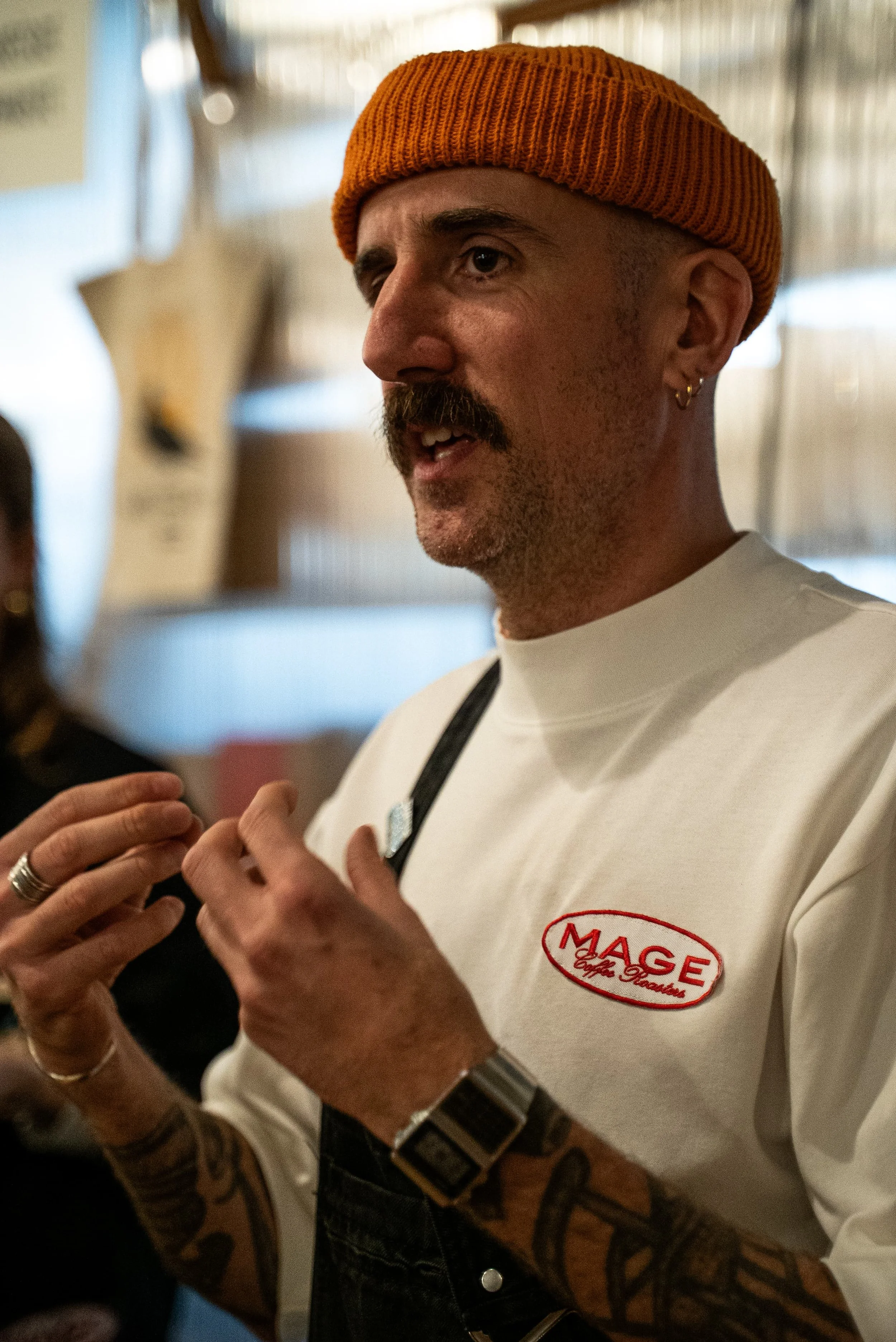 Un homme portant un bonnet orange et un t-shirt blanc avec un logo 'MAGE' en rouge, discutant avec une personne dont on voit en partie la main et un bras tatoué.