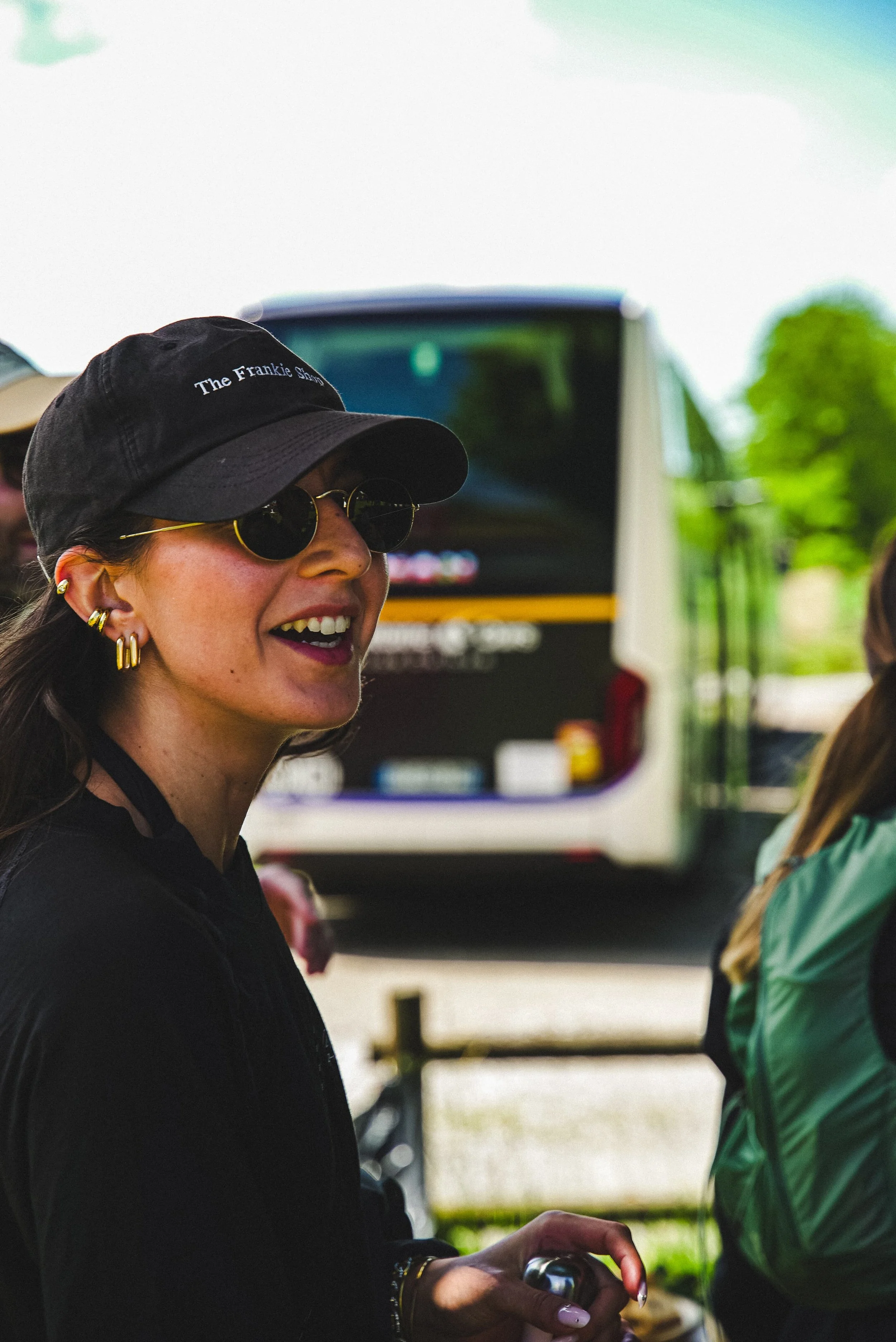 Une femme souriante portant un chapeau noir avec l'inscription 'The Frankie Show', lunettes rondes, plusieurs boucles d'oreilles en or, et une veste noire, lors d'une sortie en plein air avec un bus en arrière-plan.