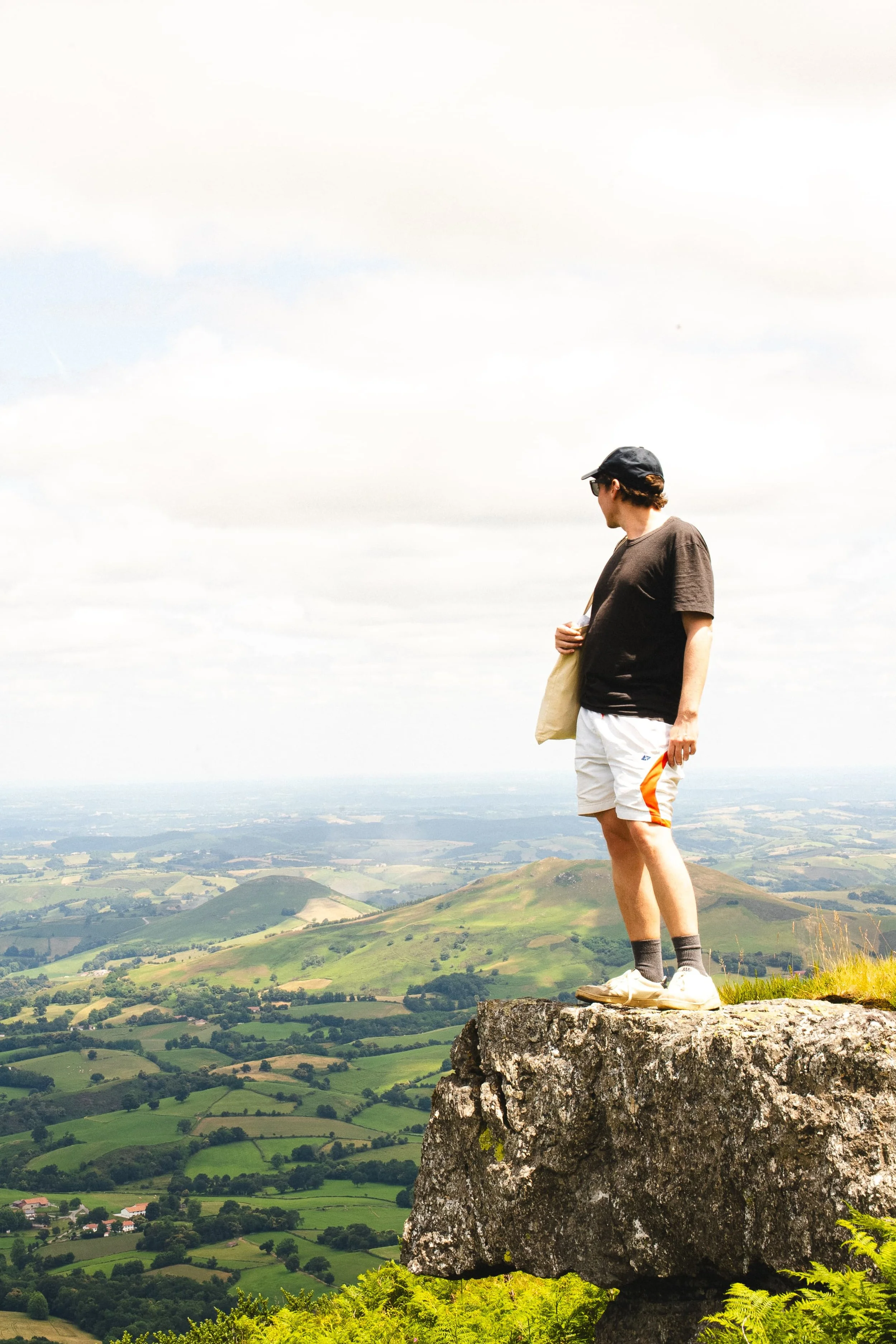 Un jeune homme debout sur une grande roche au sommet d'une colline, regardant vers la droite, avec un paysage verdoyant en contrebas, sous un ciel partiellement nuageux.