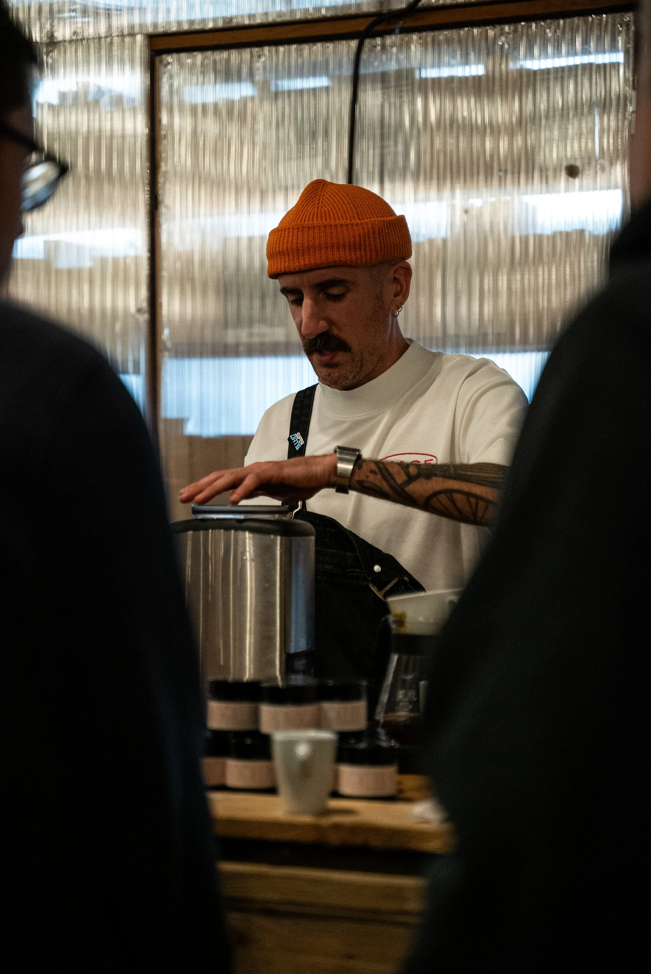 Un homme portant un bonnet orange, une chemise blanche, avec un tatouage sur le bras, en train de préparer du café dans une cuisine.