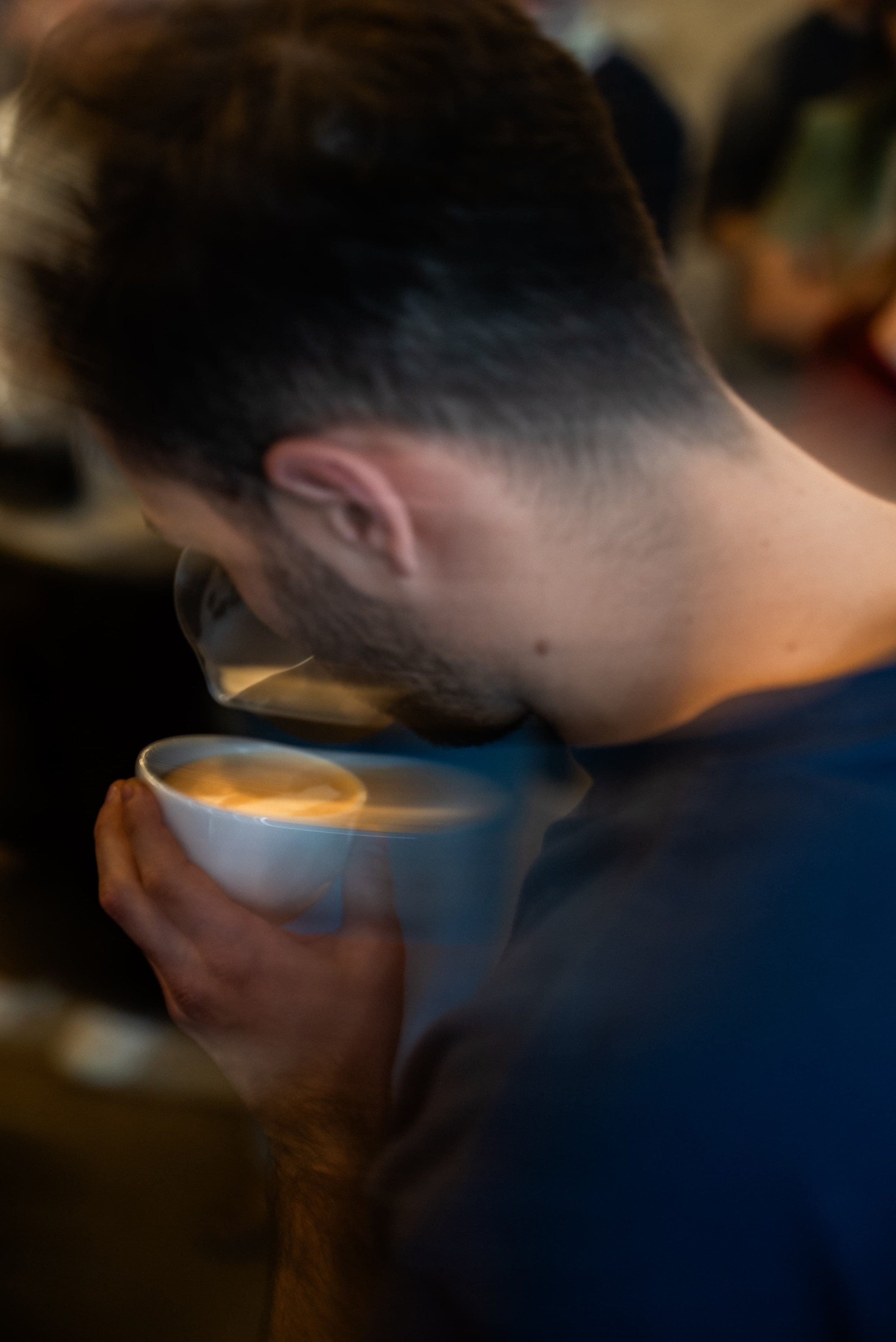 Un homme avec une coupe de cheveux courte et une barbe, il tient une tasse blanche, semble manger ou boire quelque chose dans une ambiance tamisée.