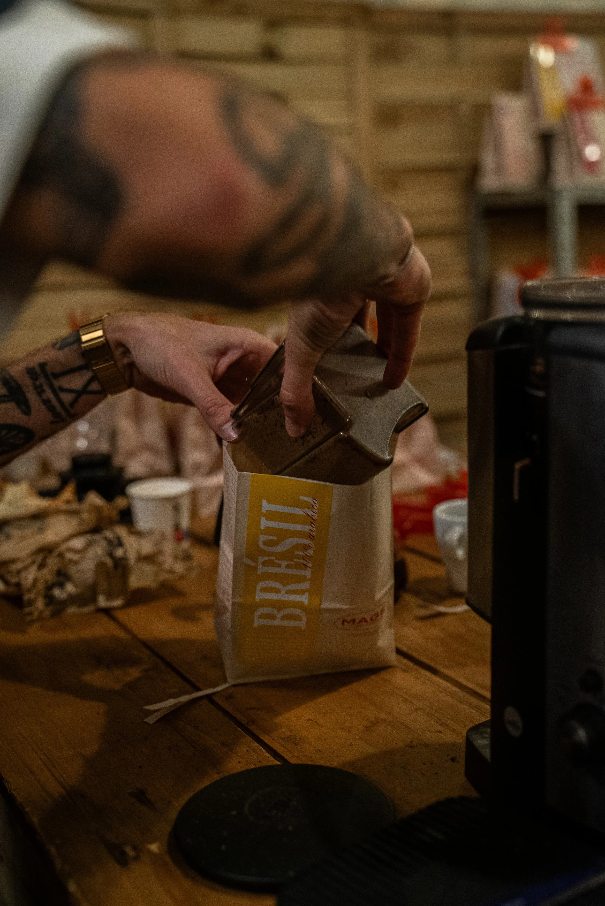 Une main de tatoué avec une montre en or verse de la farine d'une boîte en papier dans une machine à café. La scène se passe dans une cuisine en bois.