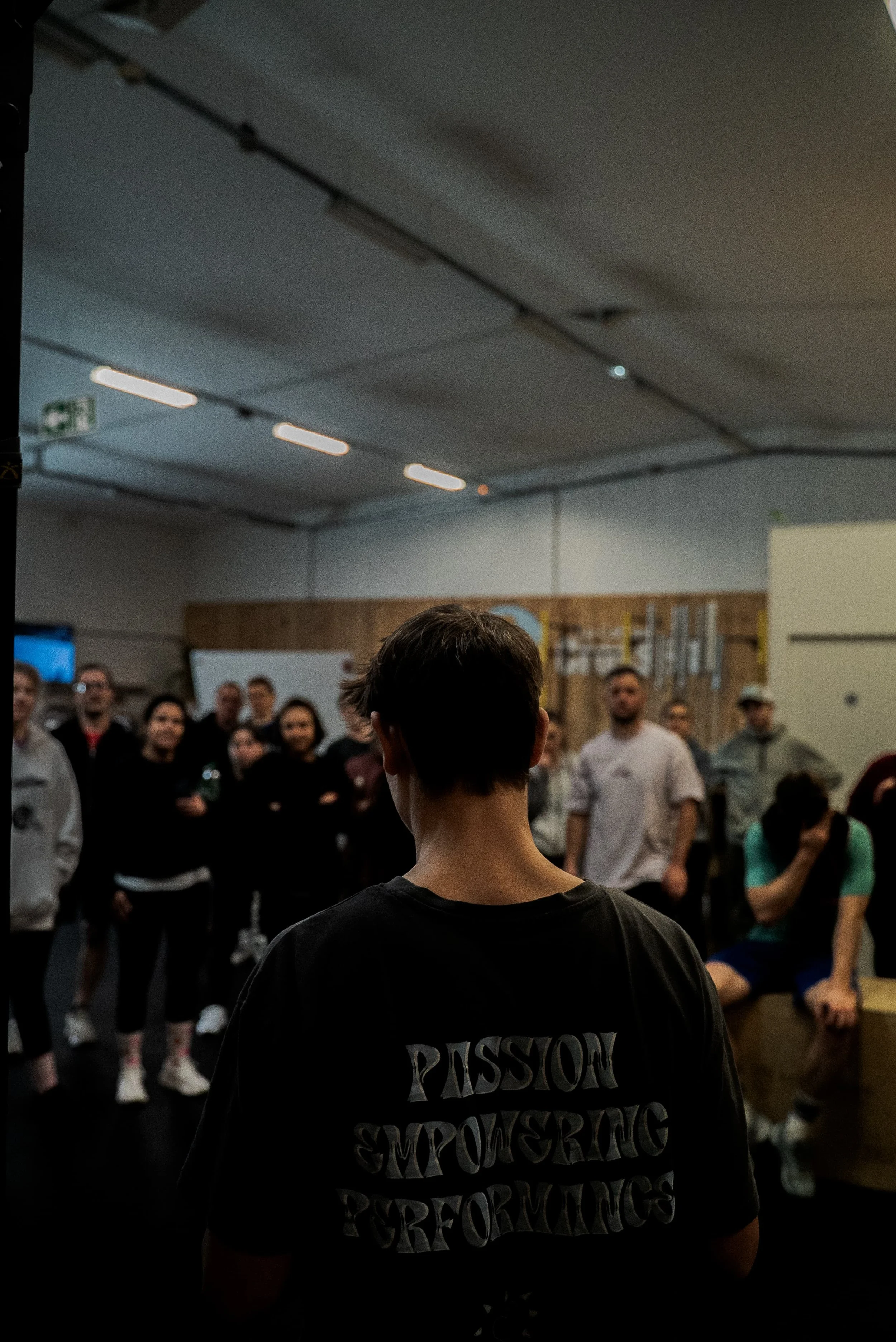 Salle avec un groupe de personnes, vue du dos d'une personne portant un t-shirt avec des mots en anglais, dans une ambiance de présentation ou réunion.