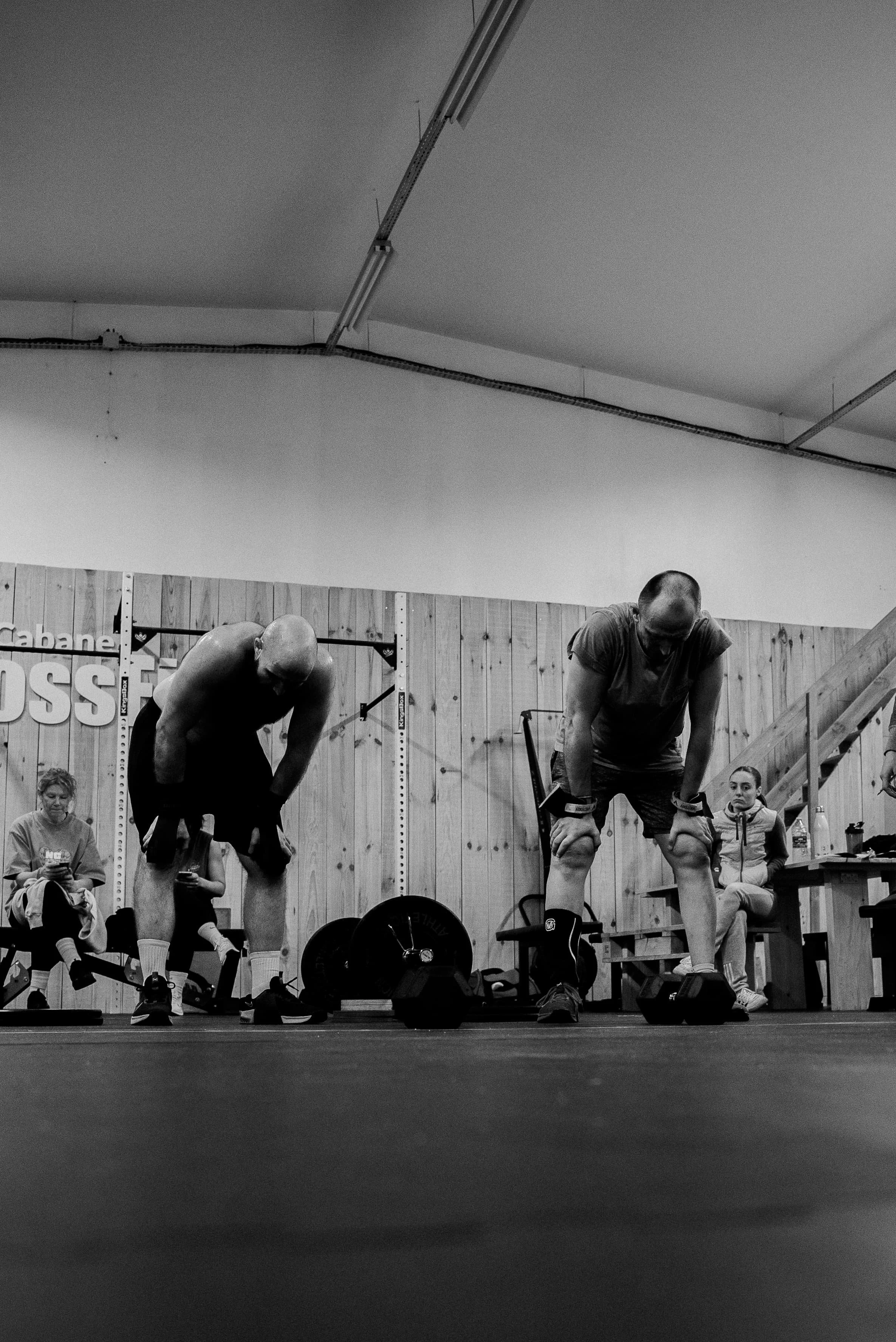 Deux hommes en train de faire des exercices de musculation dans une salle, avec plusieurs personnes assises en arrière-plan et des poids au sol.