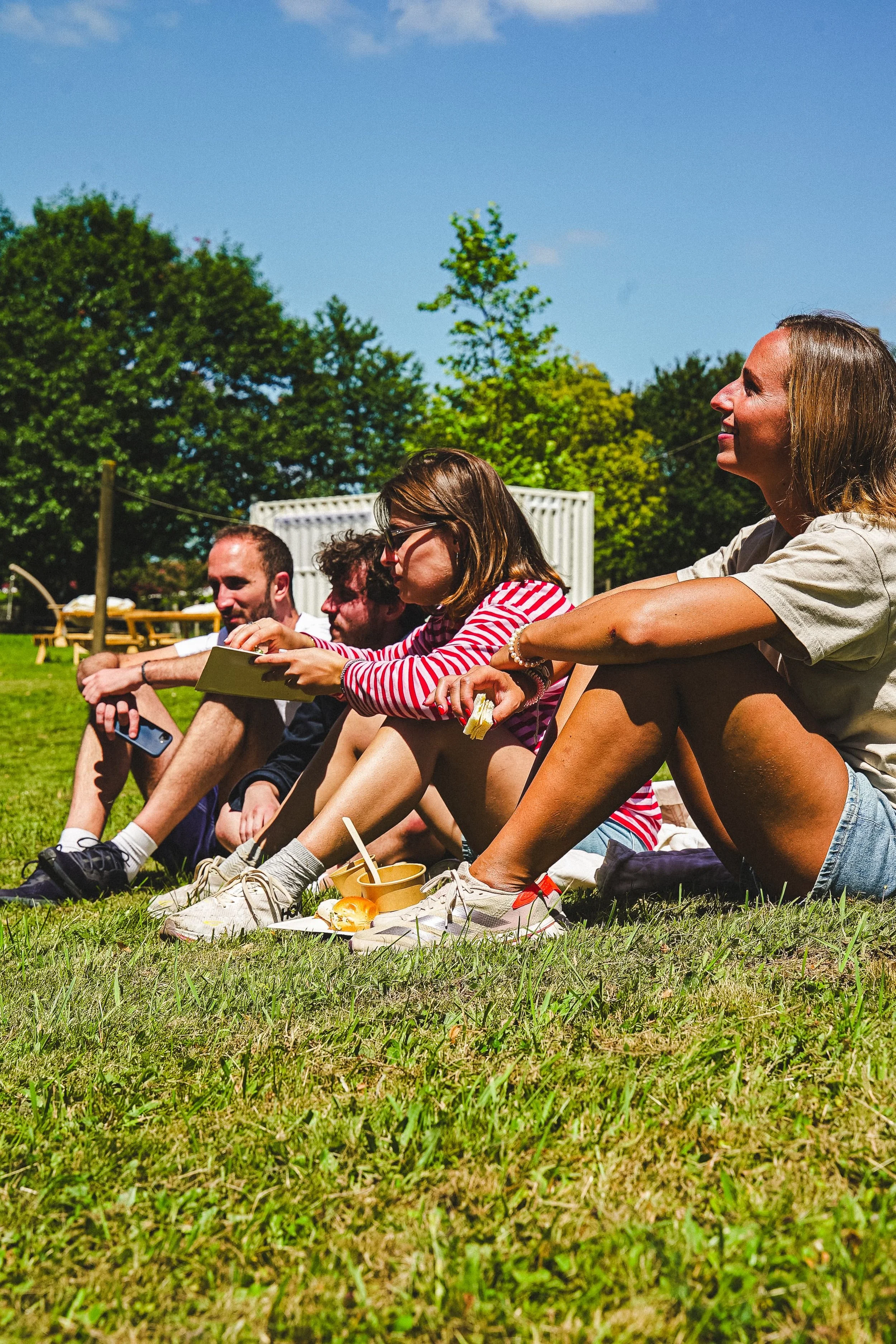Groupe de personnes assises sur l'herbe lors d'un pique-nique en plein air, avec des arbres verts en arrière-plan, un ciel bleu et des aliments devant elles.