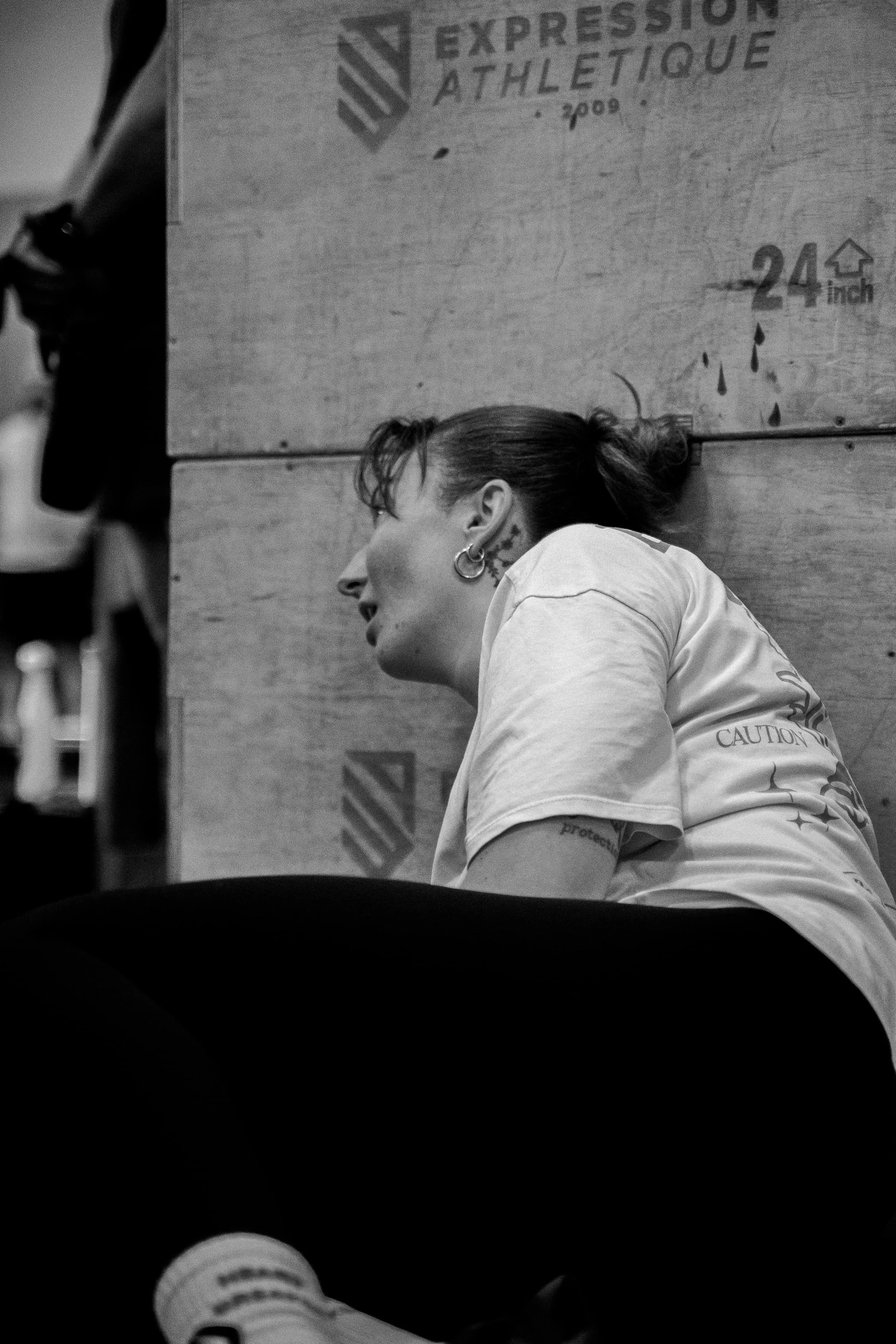 Une femme aux cheveux courts, portant des boucles d'oreilles et un t-shirt, est assise contre un mur en bois. On peut voir une boîte en carton dans le fond avec le texte 'EXPRESS ATHLETIQUE'.