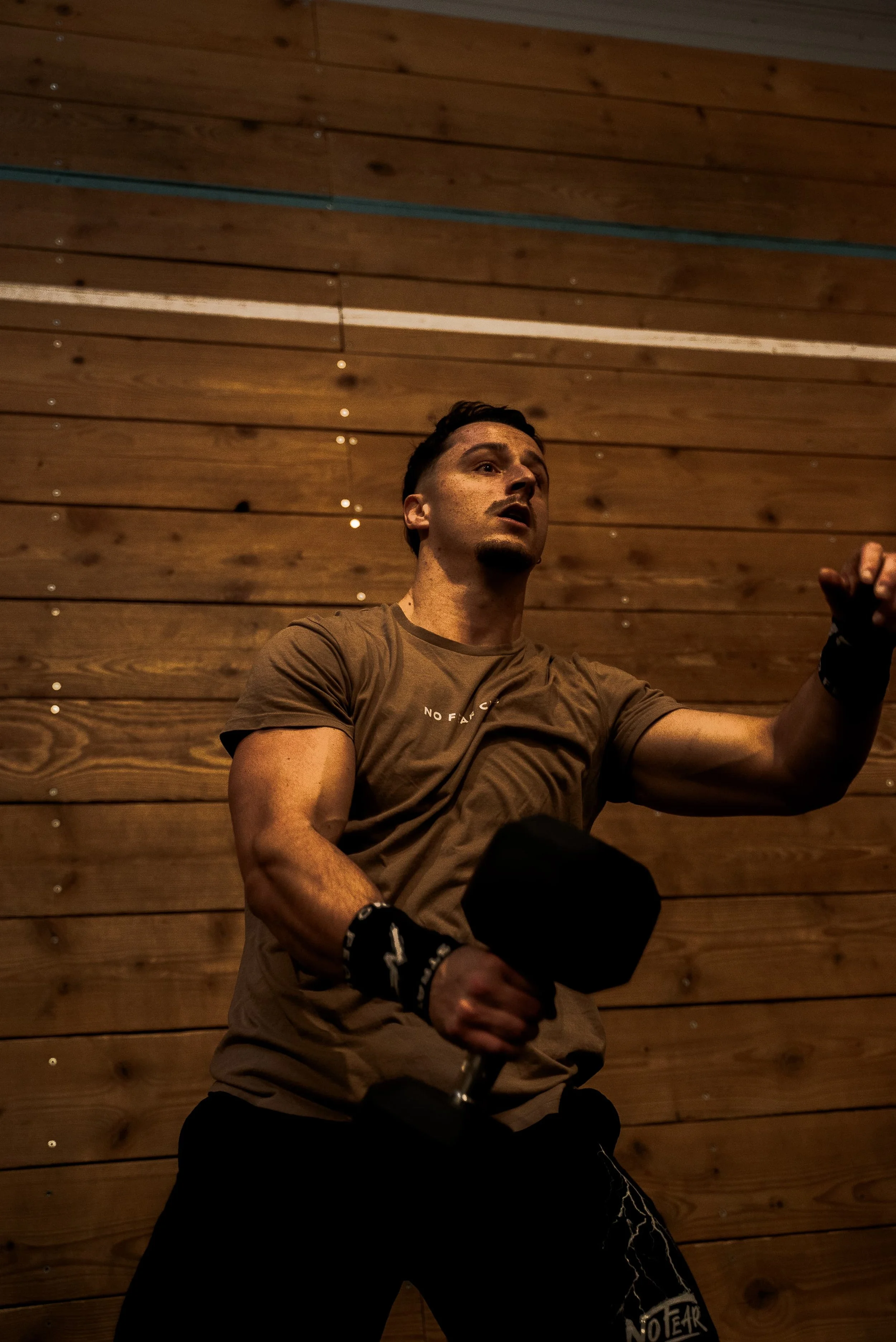 Un homme en tenue de sport, portant des gants de boxe, semble faire de l'exercice de boxe en intérieur, avec un mur en bois en arrière-plan.