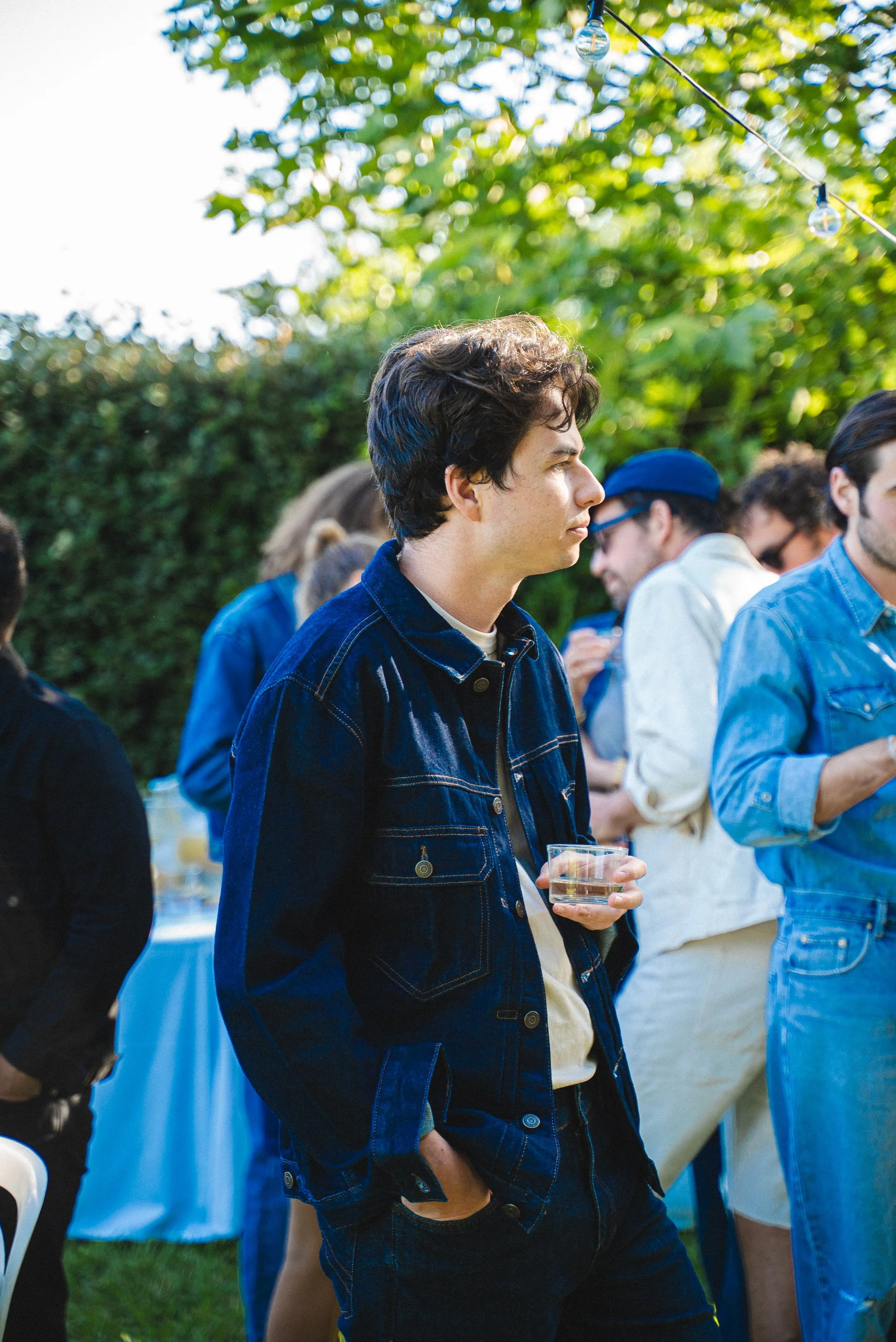 Jeune homme avec cheveux bruns, portant chemise en denim, tenant un verre à cocktail, lors d'un événement en plein air avec d'autres personnes en fond.