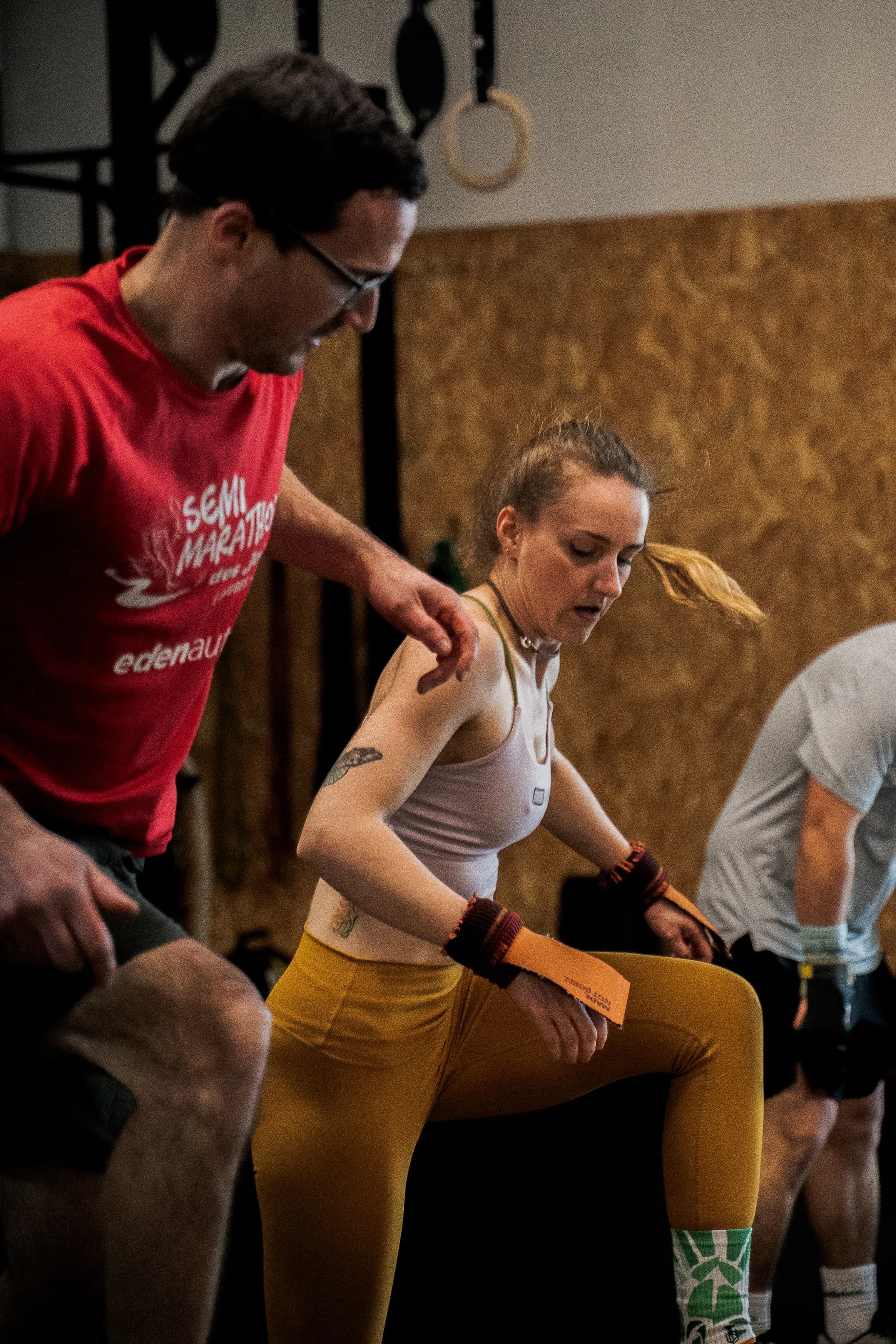 Une femme assise en train de faire du sport, accompagnée d'un homme debout à côté d'elle dans un environnement intérieur.