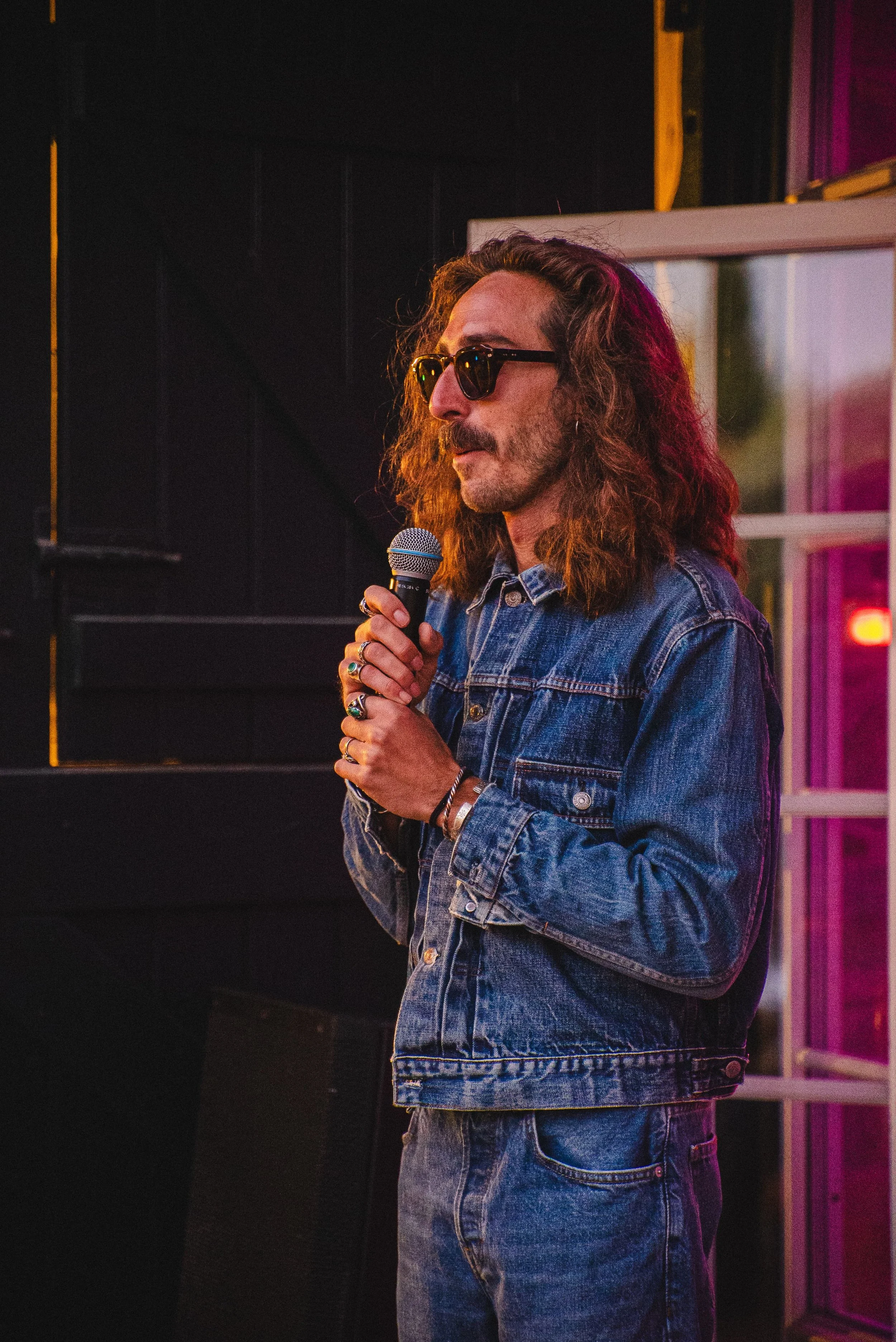 Un homme portant des lunettes de soleil, une veste en denim, tenant un microphone, dans un environnement avec un éclairage coloré