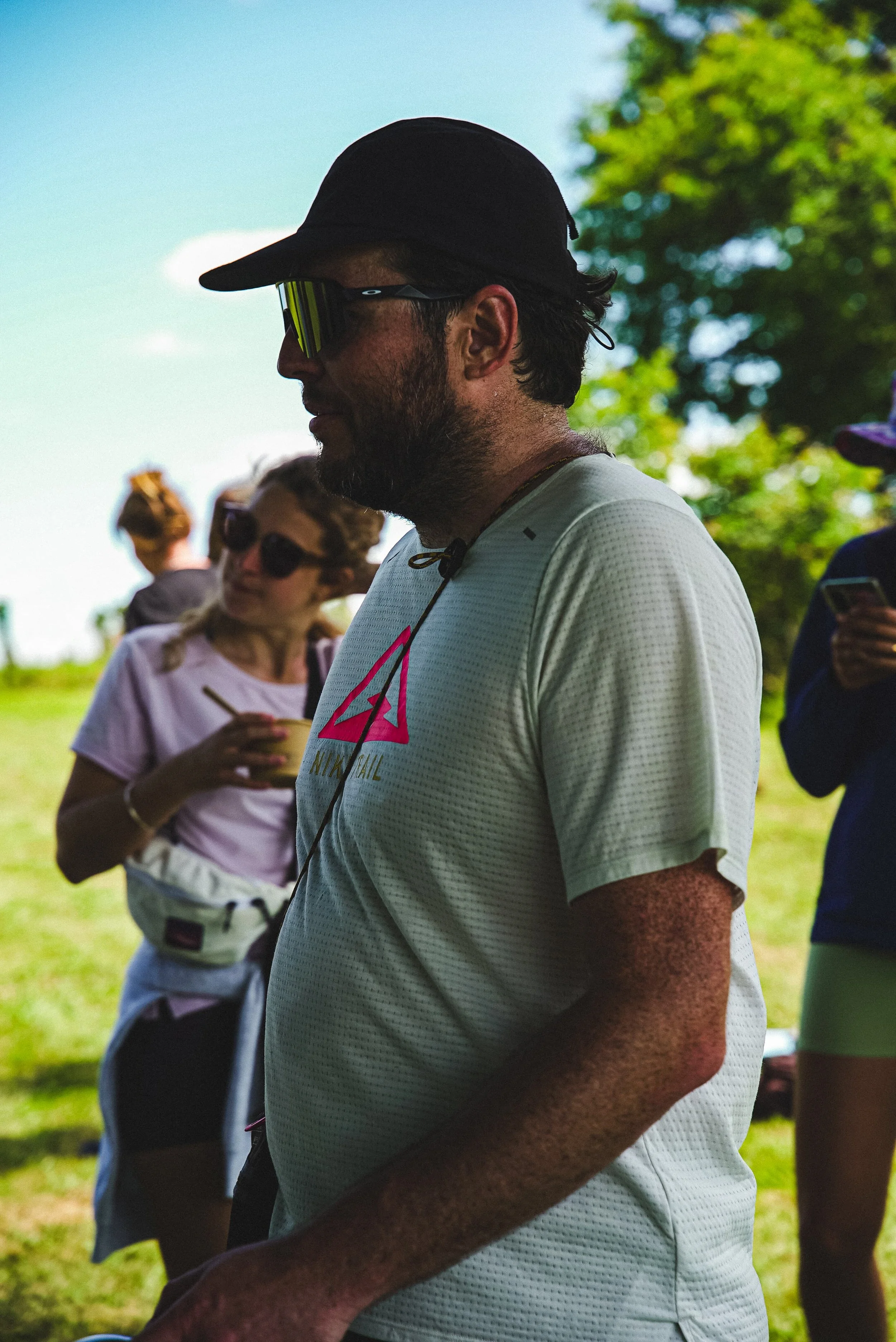 Homme portant une casquette, des lunettes de soleil et un t-shirt blanc, s'air concentré lors d'une activité extérieure par une journée ensoleillée avec des arbres en arrière-plan.