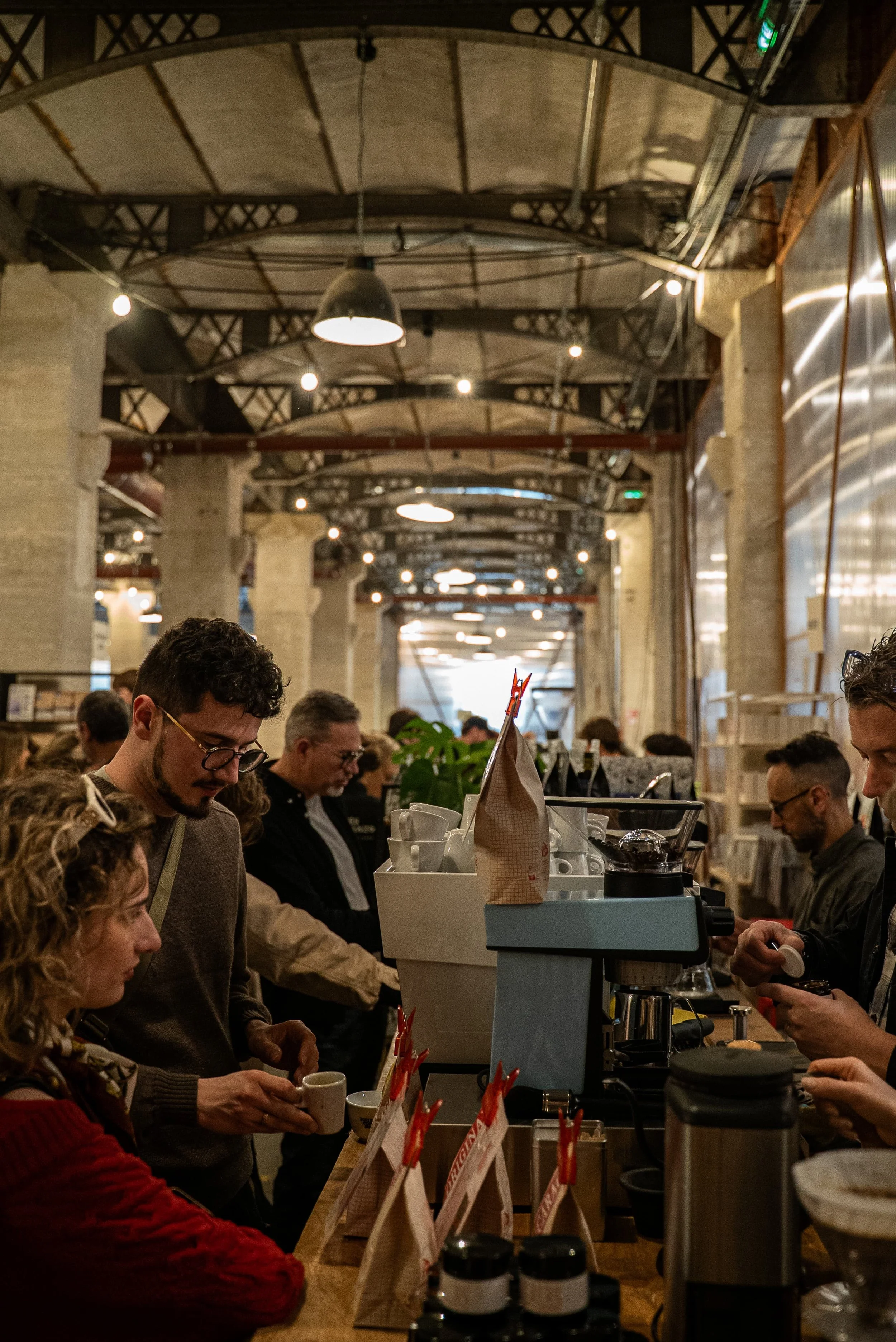 Gens faisant la queue au comptoir d'un café ou d'un restaurant dans un espace industriel avec des poutres en acier et des murs en briques, ambiance chaleureuse et lumineuse.
