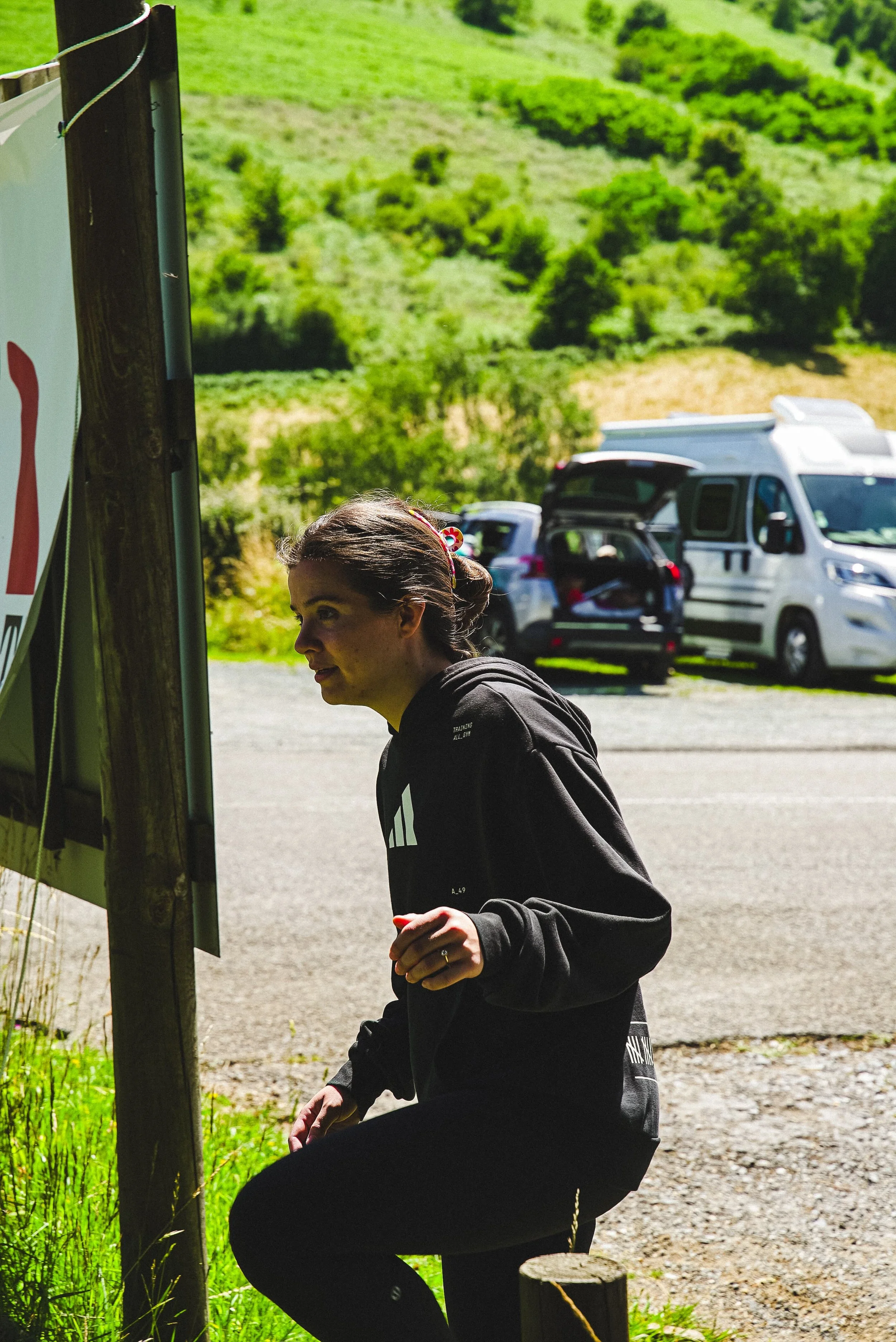 Une femme avec des cheveux bruns attachés, portant un sweat noir avec un logo blanc, est assise ou accroupie près d’un panneau ou d’un affichage en plein air, avec des véhicules en arrière-plan et un paysage verdoyant.