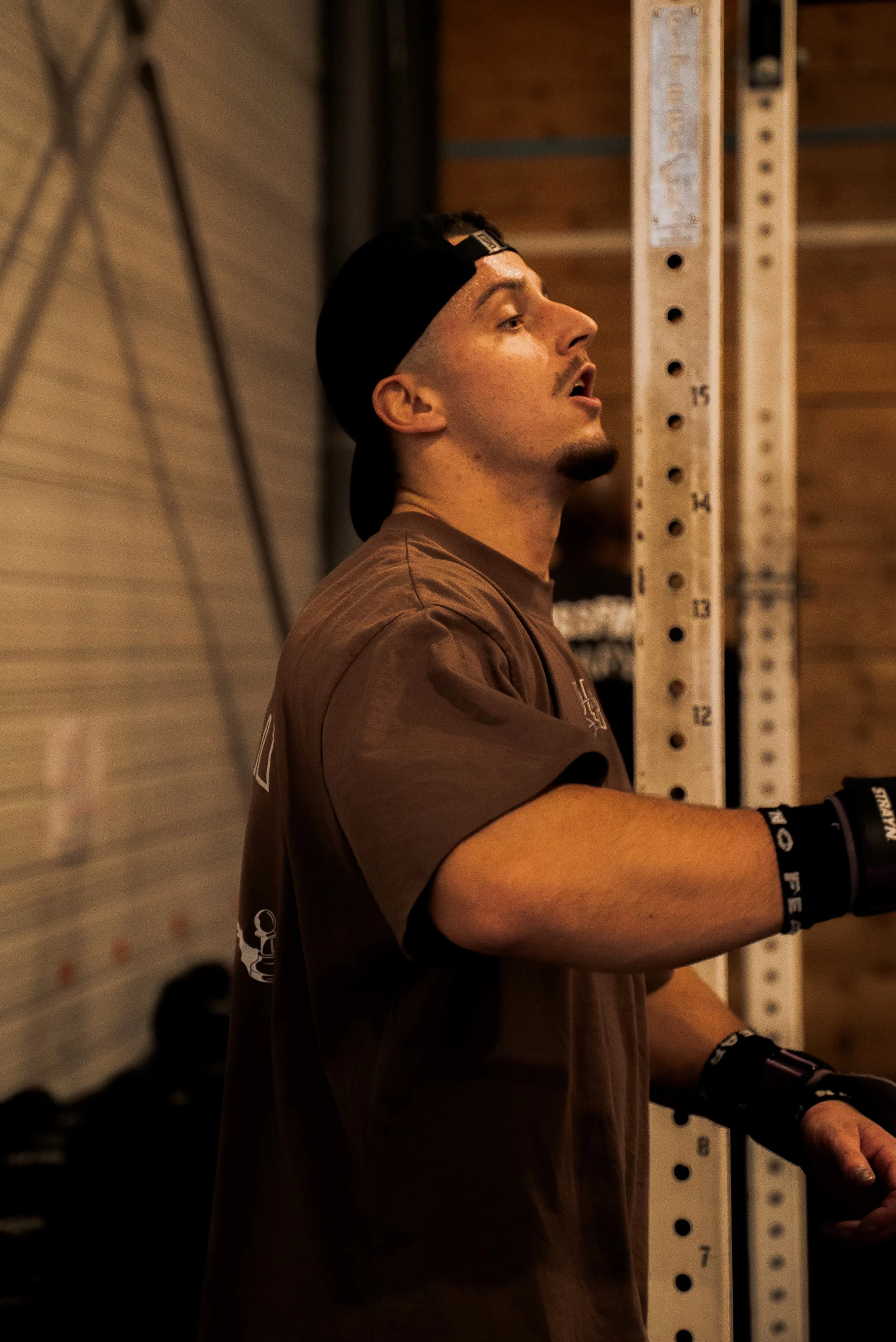 Un homme portant un t-shirt brun, un casque noir à l'envers, utilise des poids de musculation dans une salle de sport.