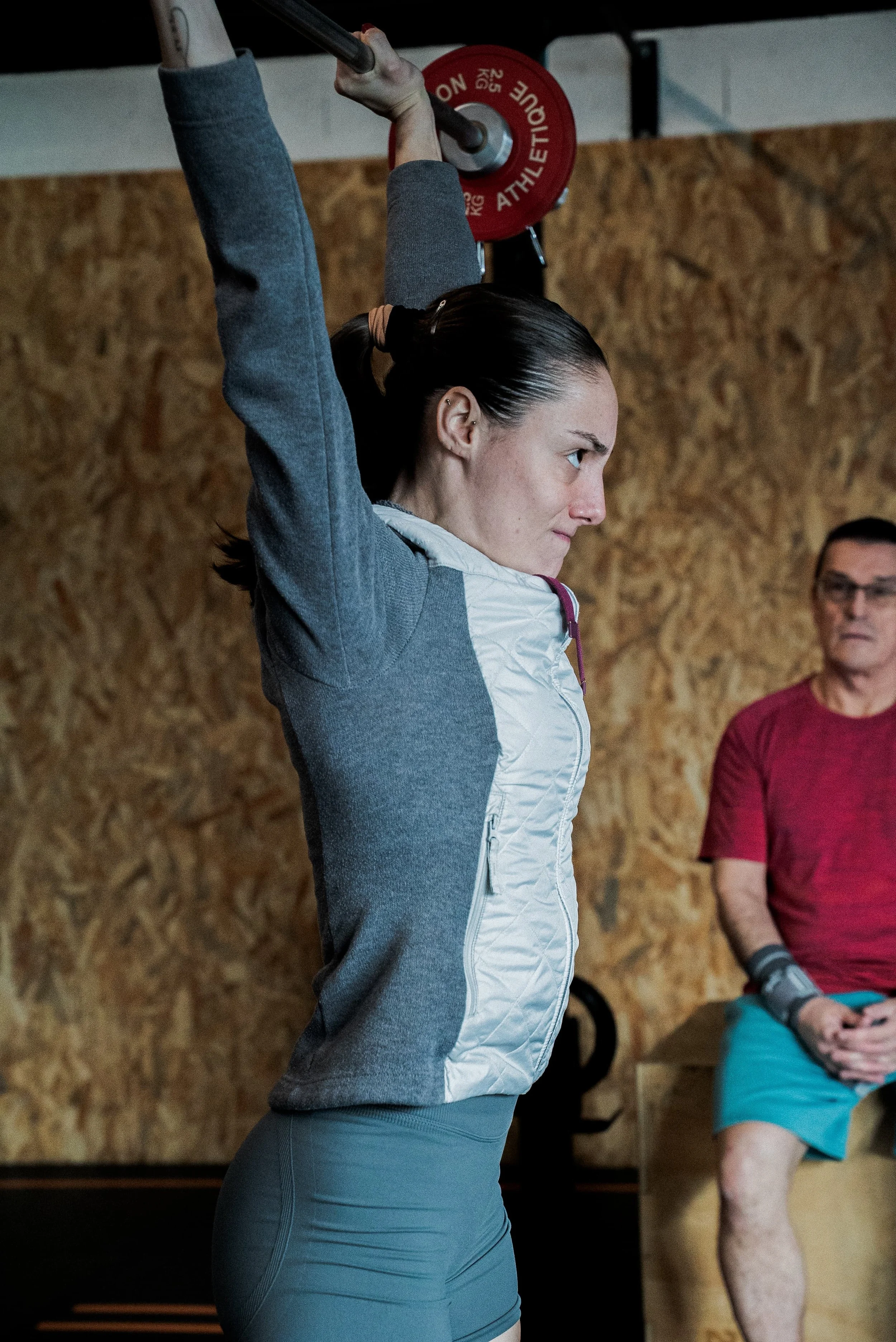 Une femme soulève une barre avec un poids rouge à l'entraînement, portant une veste grise et un haut de sport gris. À l'arrière, un homme en t-shirt rouge observe, dans une salle de sport avec un mur en bois.