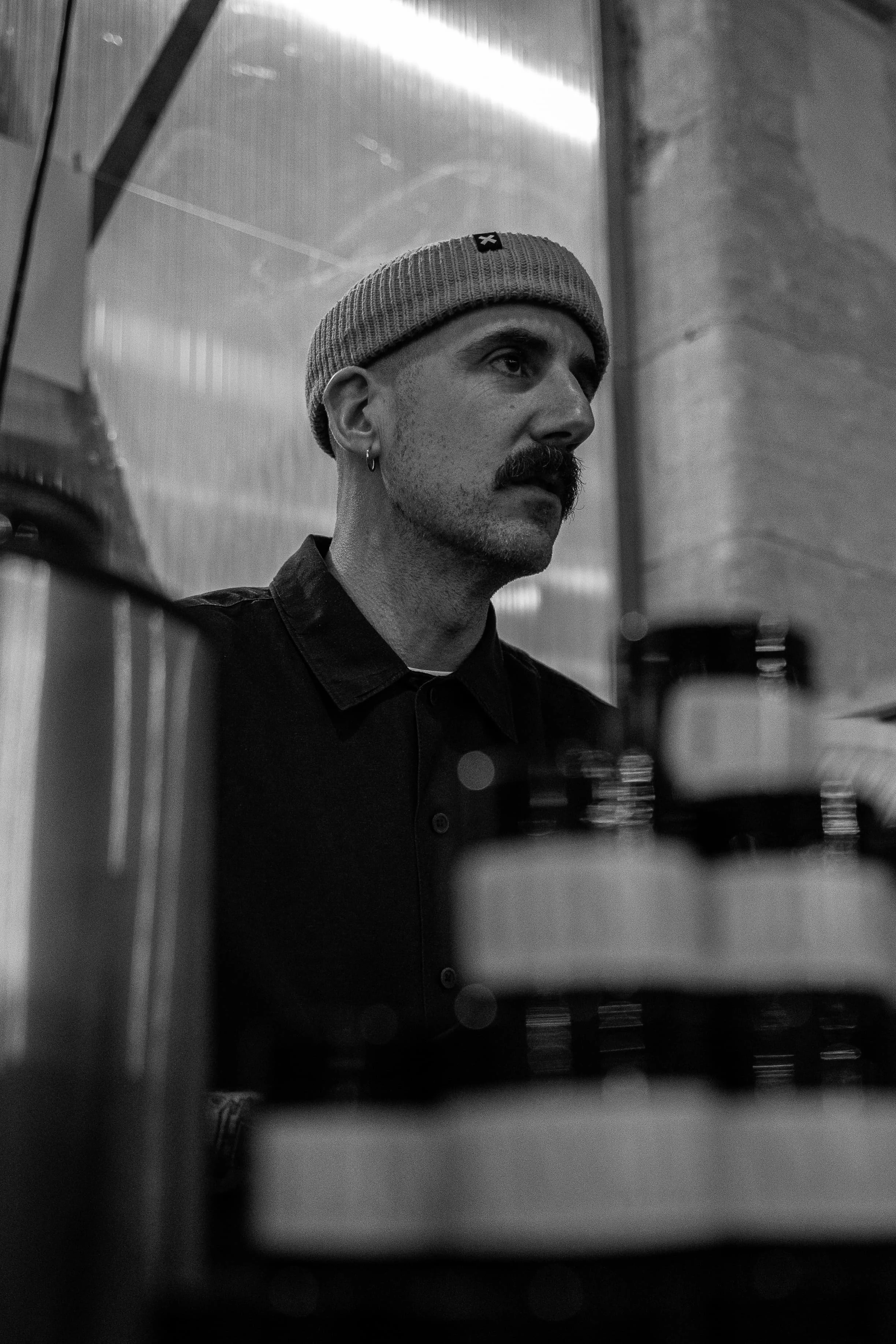 Un homme avec une moustache et des boucles d'oreilles porte un bonnet gris et une veste noire, regardant vers la droite, dans un espace intérieur en noir et blanc.