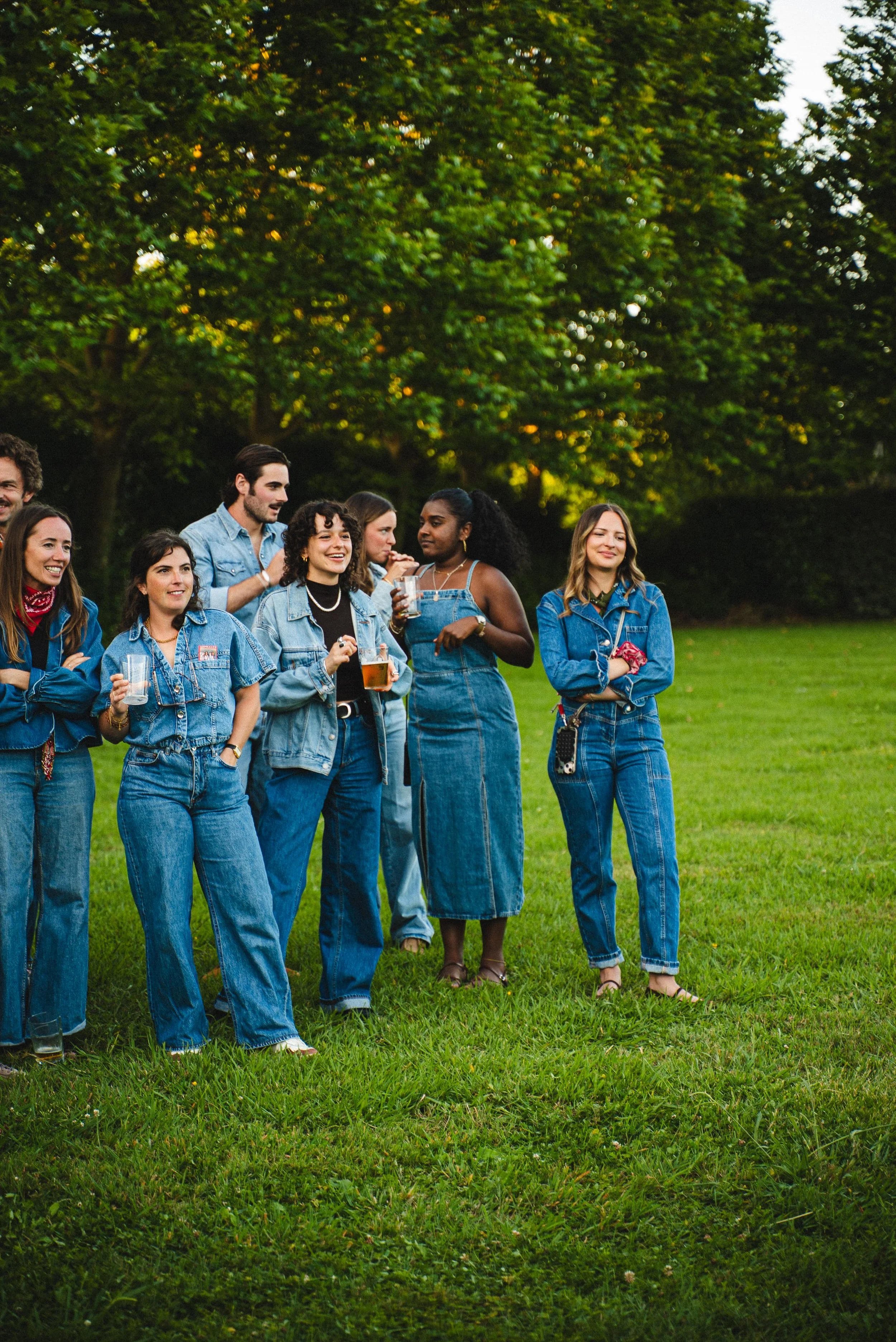 Groupe de jeunes adultes portant des vêtements en jean, se tenant en cercle dans un parc vert, certains avec des boissons à la main.