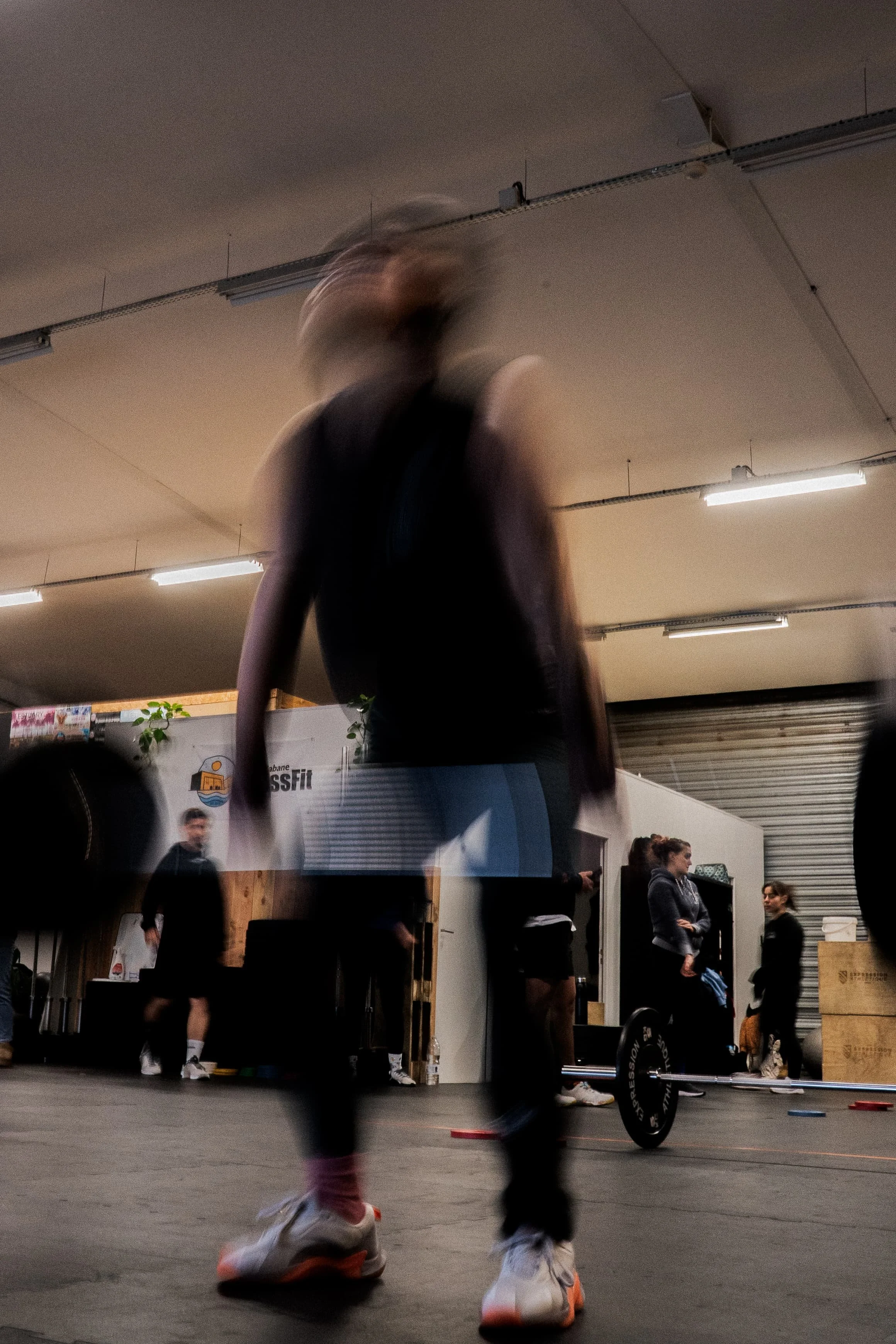 Une personne en train de faire de la musculation dans une salle de sport, avec d'autres personnes en arrière-plan. La personne porte des vêtements de sport et des chaussures blanches avec une semelle orange.