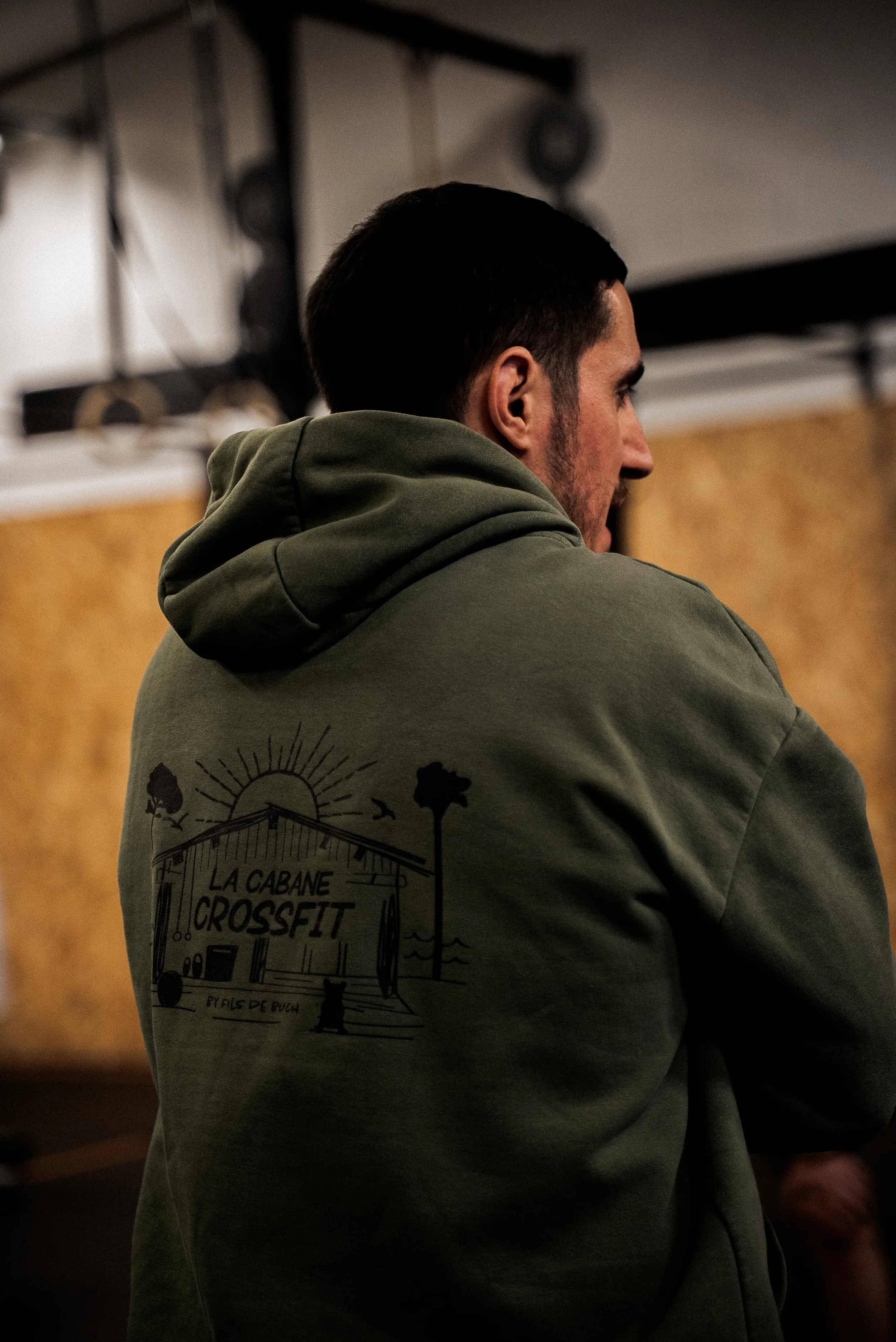 Un homme vu de profil portant un sweat-shirt gris avec un logo de cabane en bois, arbres et texte 'La Cabane Crossttt'.