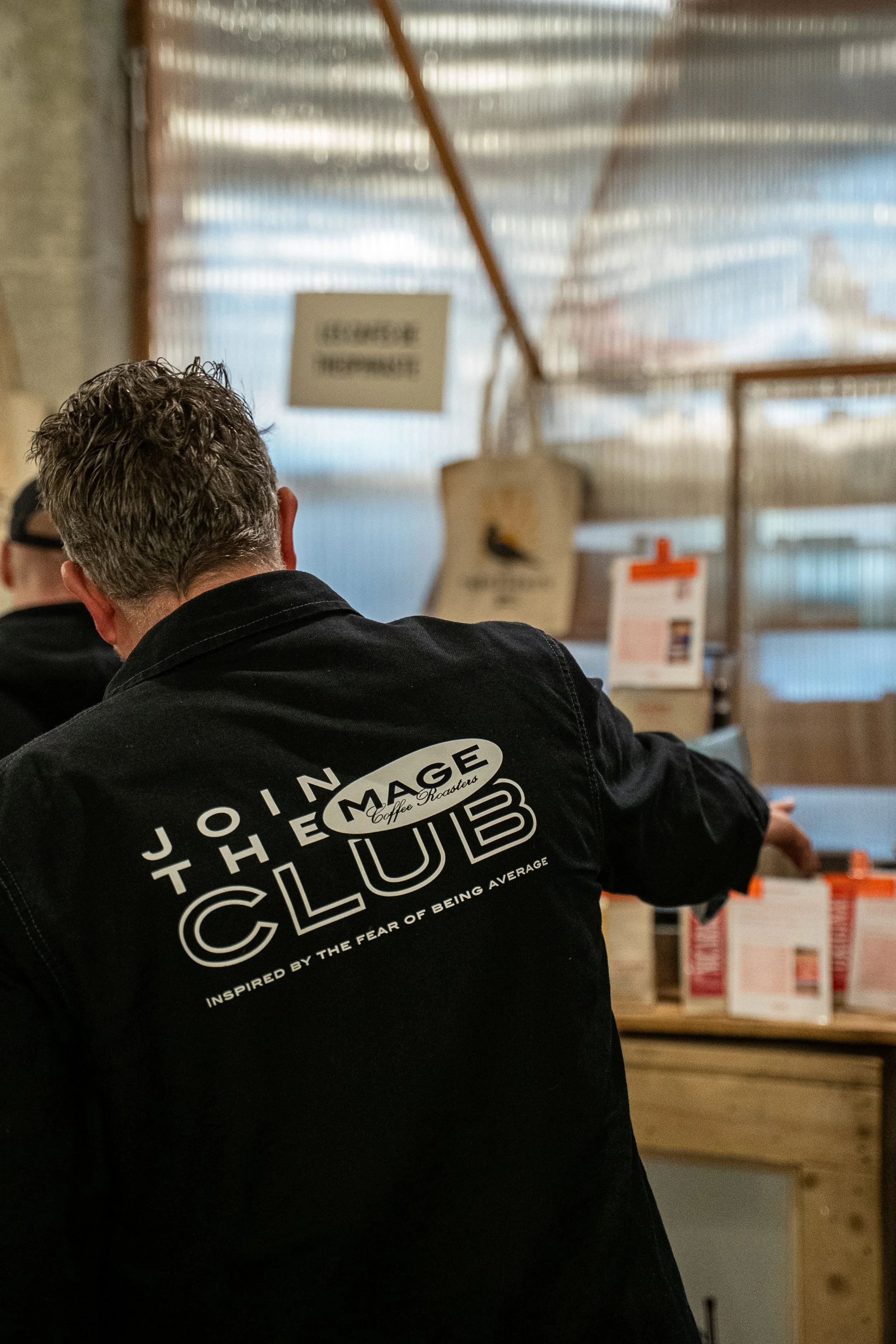 Personne portant un sweat-shirt noir avec un logo, assise à une table dans un café ou un restaurant, avec des emballages de nourriture en arrière-plan et un environnement intérieur industriel.
