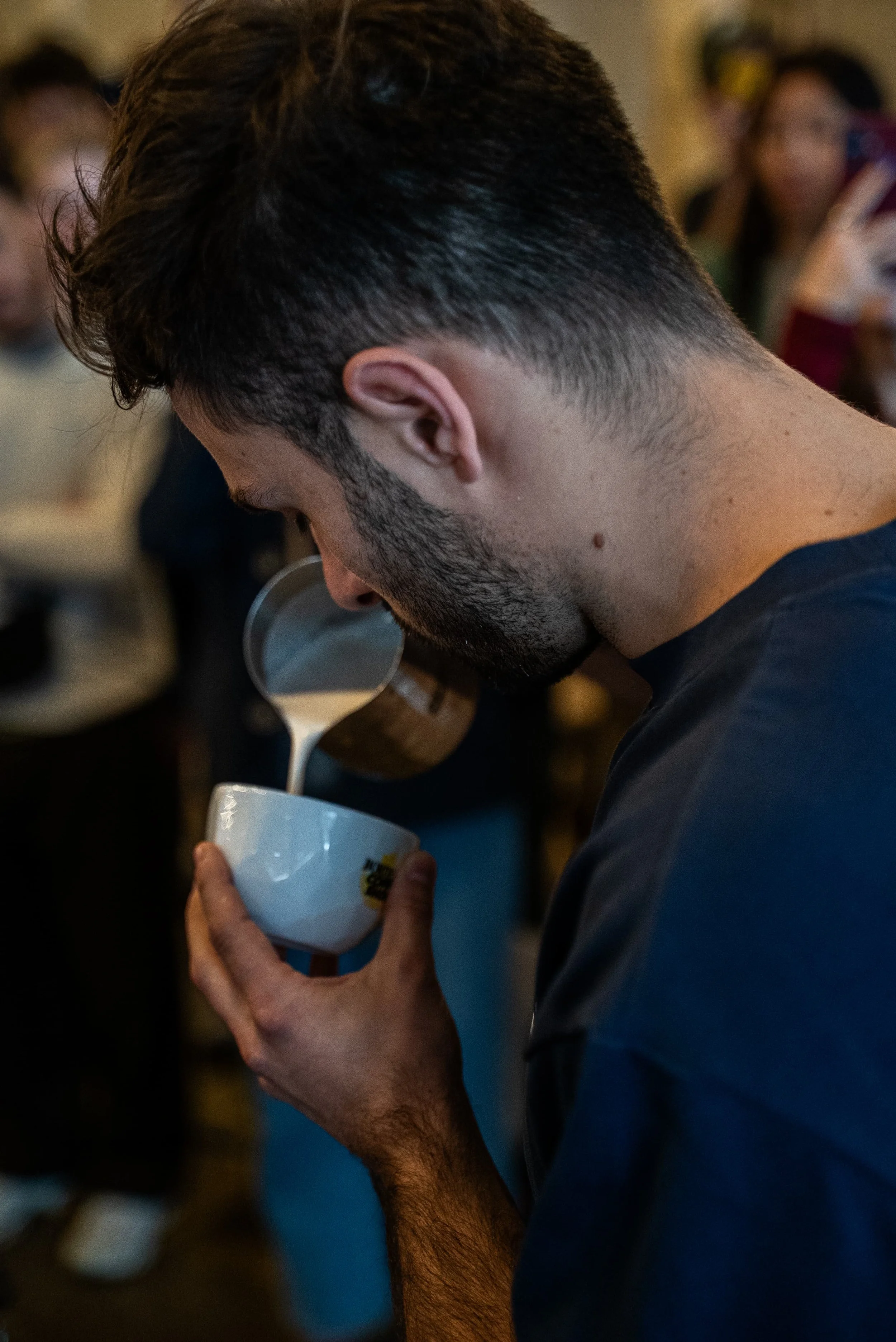 Un jeune homme avec des cheveux courts ornementés, une barbe et une moustache, qui tient un bol blanc dans une main et verse du lait dedans avec une cuillère dans l'autre main, dans une pièce où d'autres personnes sont visibles en arrière-plan.