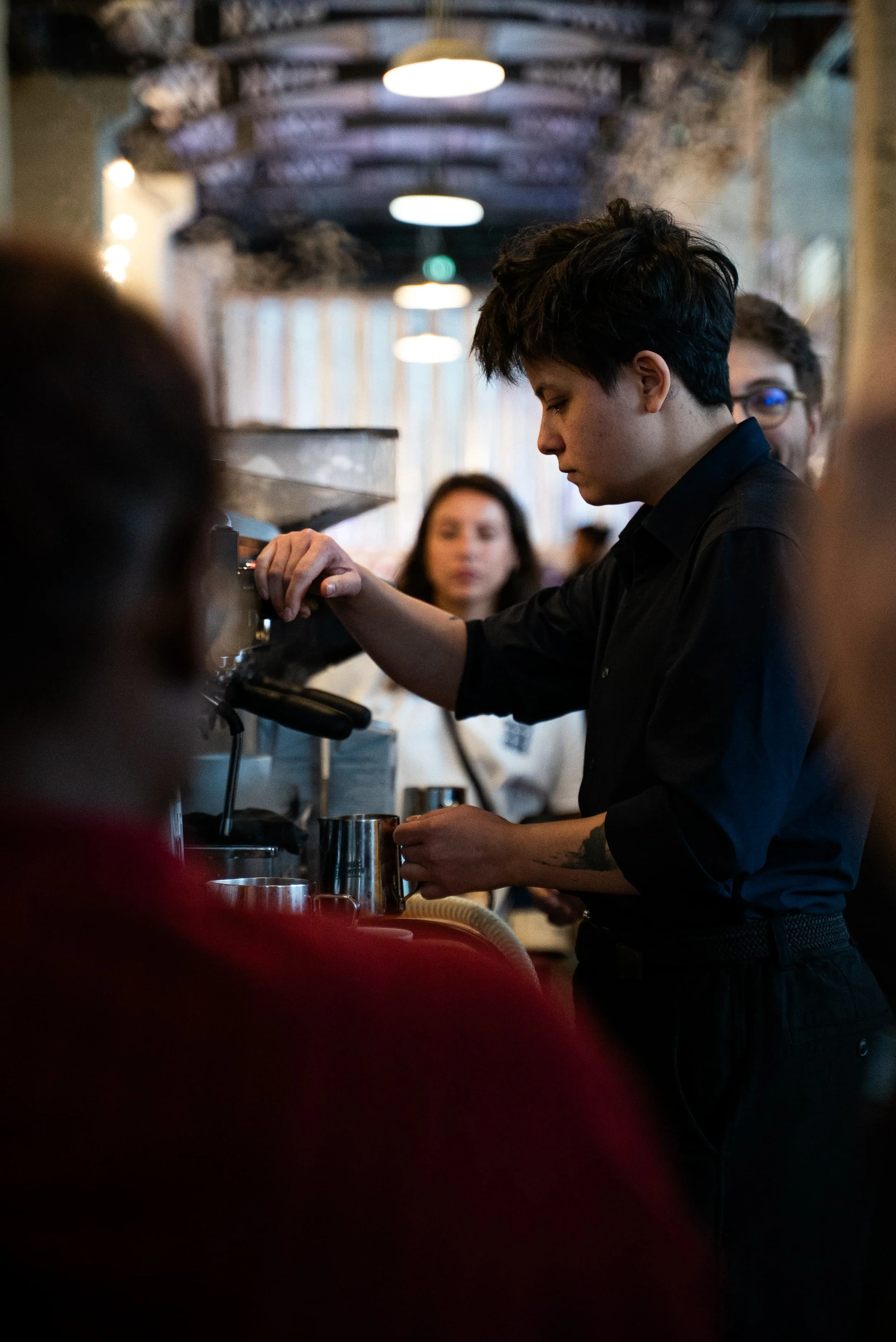 Un jeune homme en noir prépare une boisson dans un café ou un bar, avec d'autres personnes en arrière-plan dans une ambiance chaleureuse et tamisée.