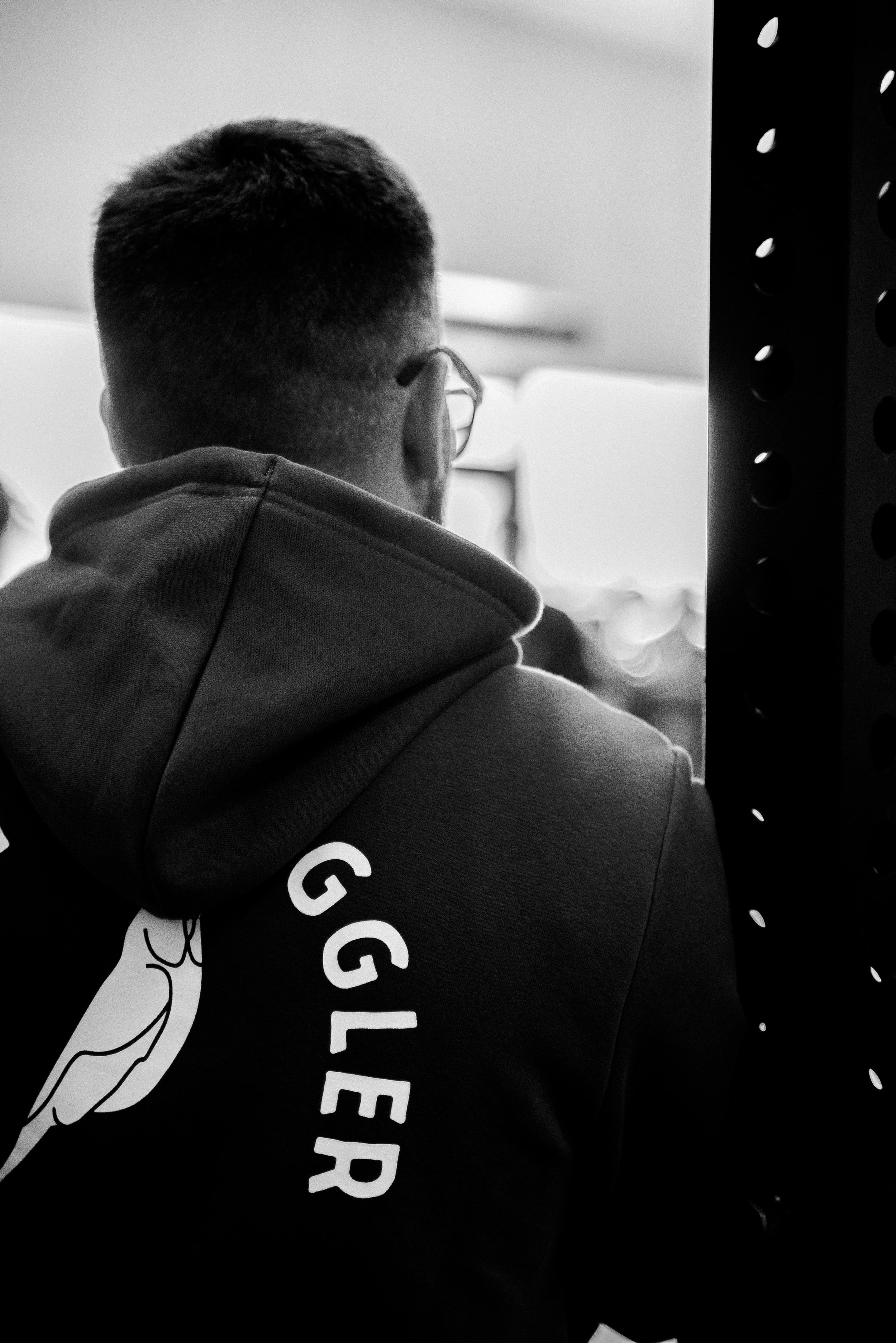 Un homme vu de dos portant un sweat à capuche gris avec un logo ou texte blanc, et portant des lunettes, dans un environnement intérieur.