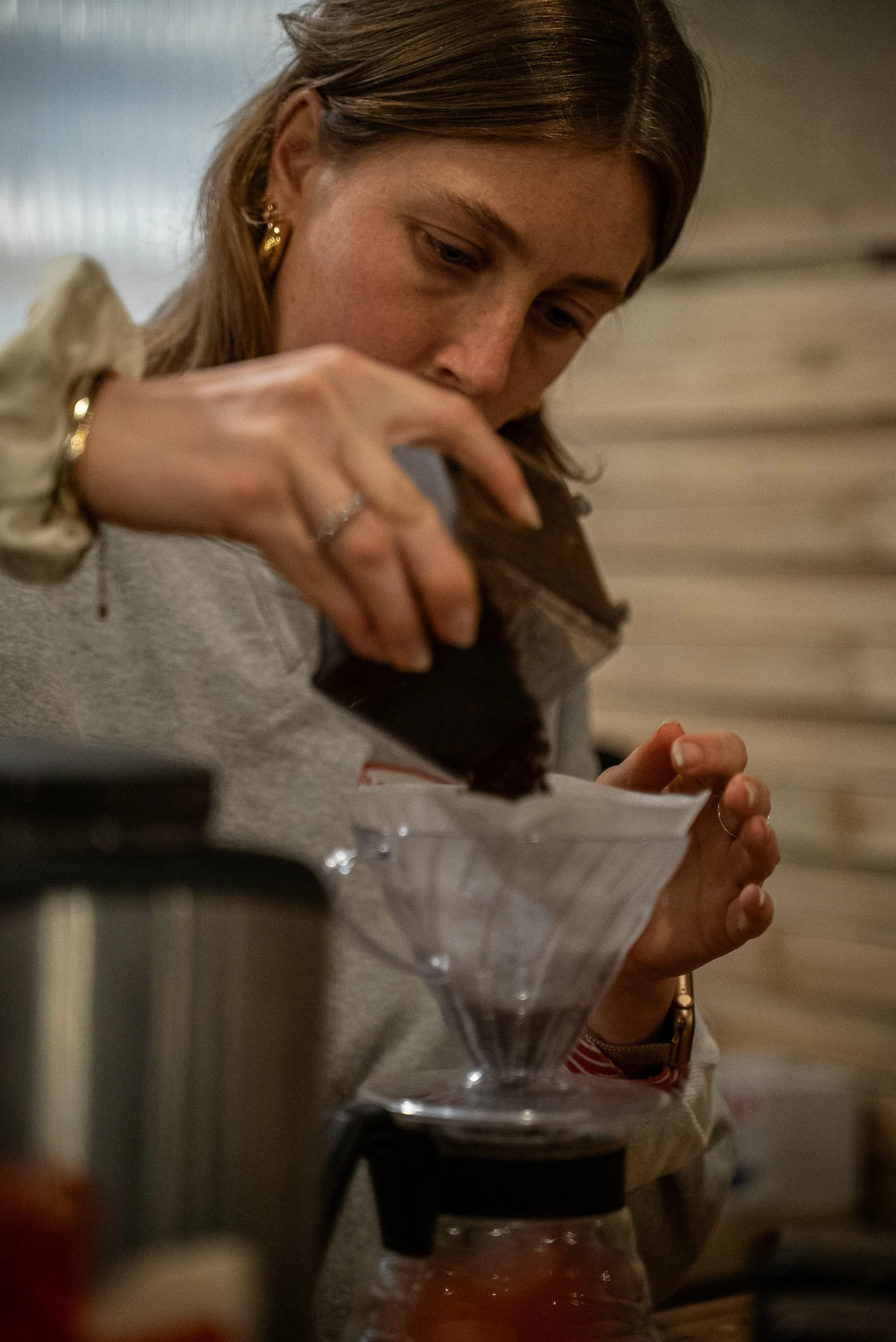 Une femme avec des boucles d'oreilles en or, qui semble concentrée, regarde un objet qu'elle tient dans la main, à proximité d'un récipient en verre.