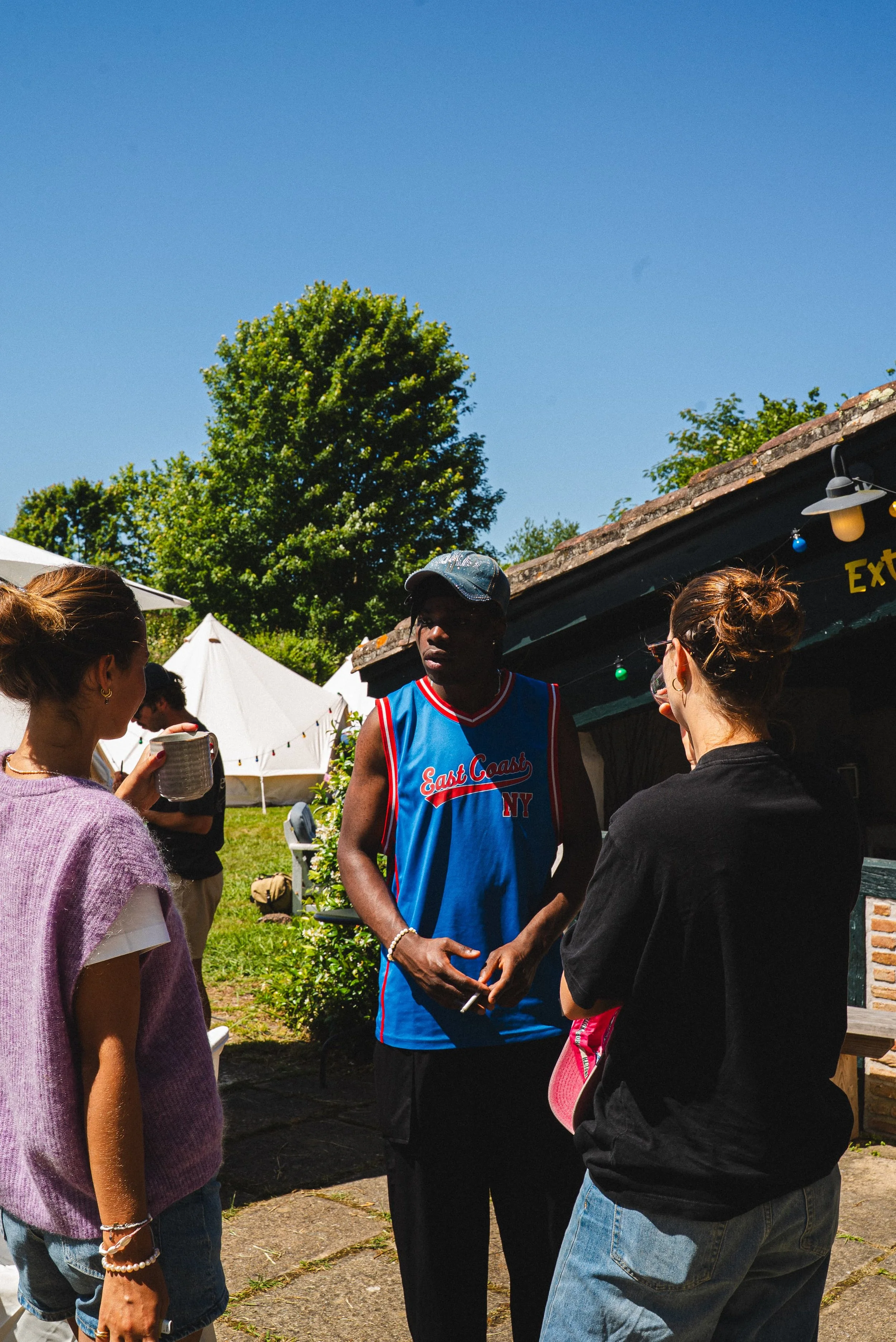 Trois personnes discutent lors d'un rassemblement extérieur par une belle journée ensoleillée, avec des parasols et des arbres en arrière-plan.