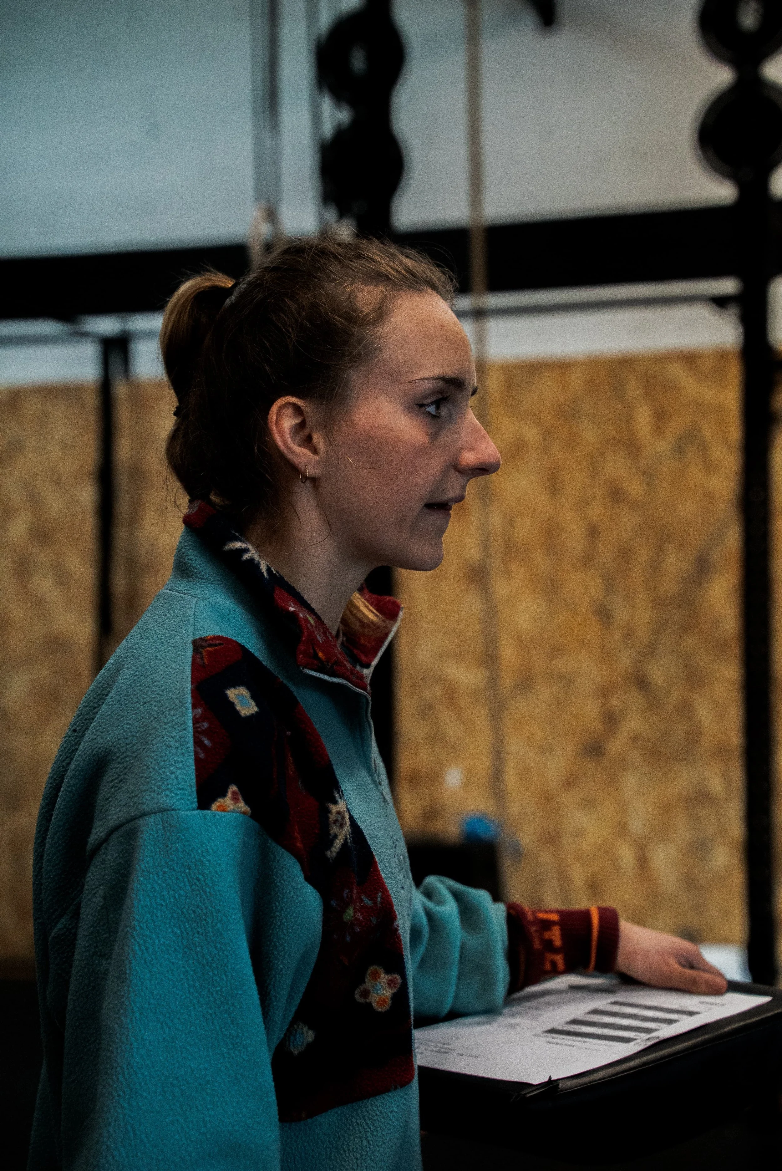 Une jeune femme avec une chemise à motifs et un pull bleu, regardant vers la droite, à l'intérieur, tenant un document avec un diagramme ou graphique et une main reposant dessus, fond en bois et structure noire.