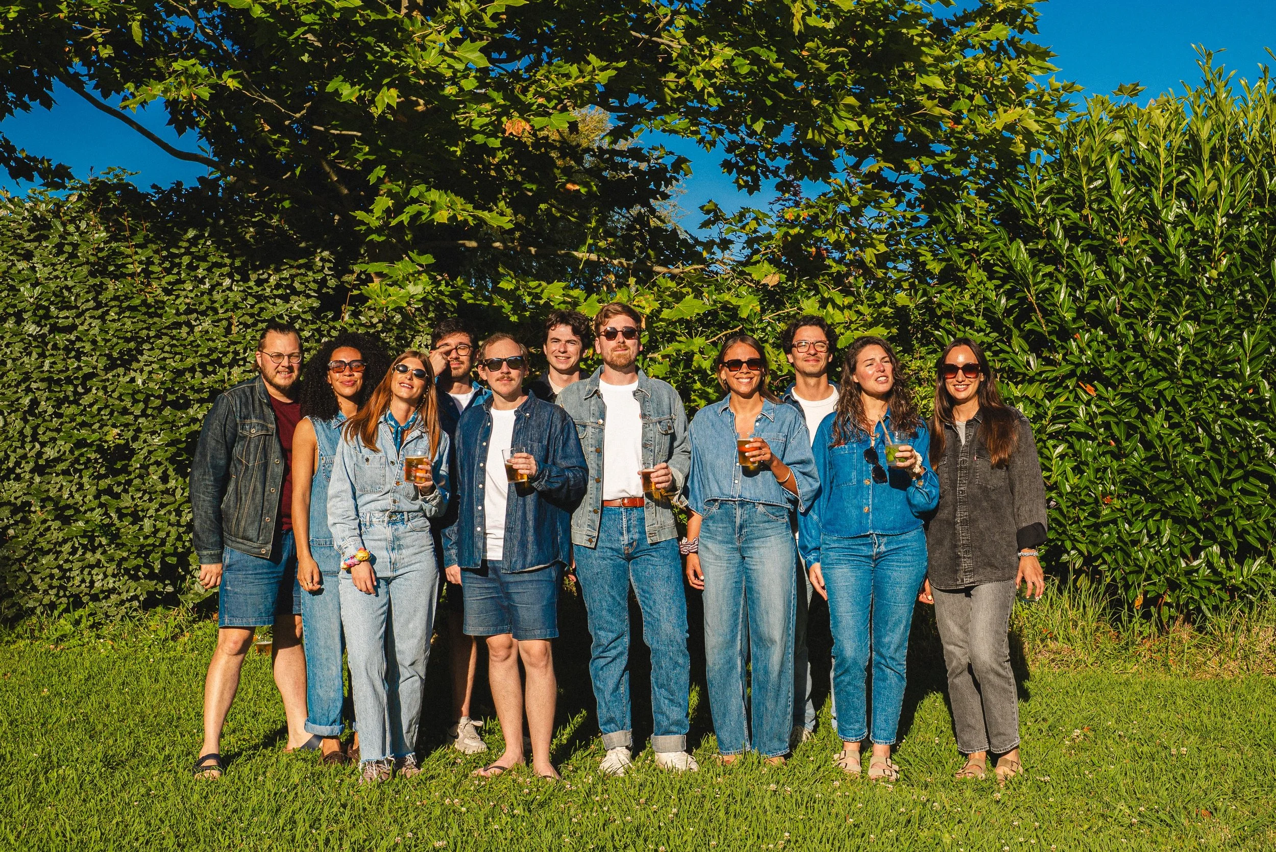 Groupe de jeunes adultes souriants, debout sur une pelouse en été, tenant des boissons, sous un ciel bleu avec des arbres verts en arrière-plan.