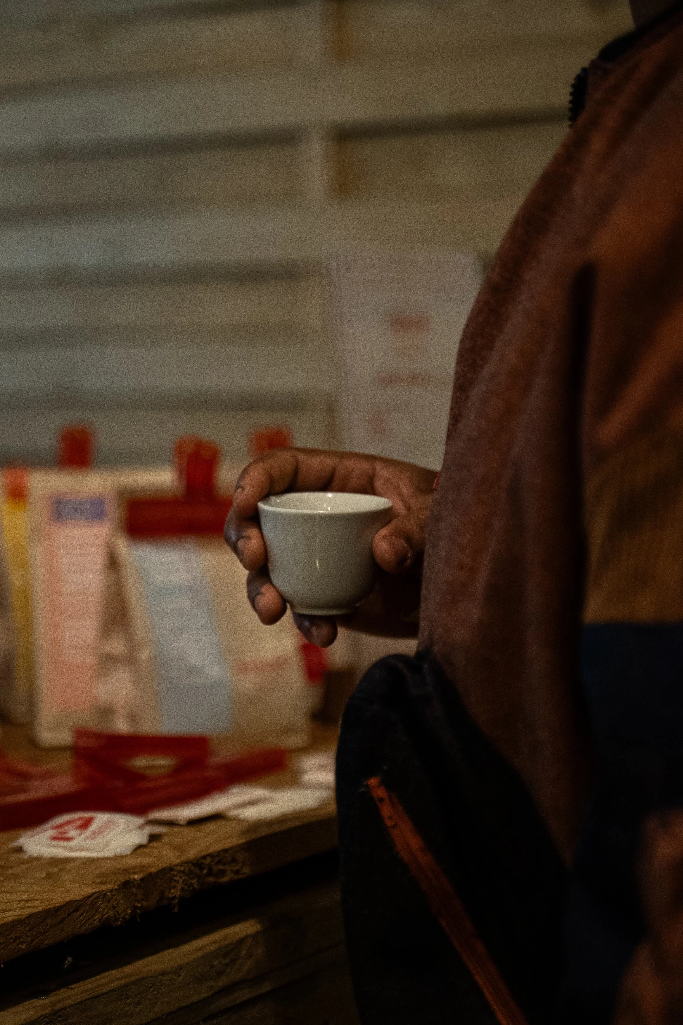 Une personne tient une petite tasse blanche avec une mainsale, vêtue d'un manteau brun, dans une pièce lumineuse. Des étagères ou un mur avec des affiches ou des objets flous en arrière-plan.