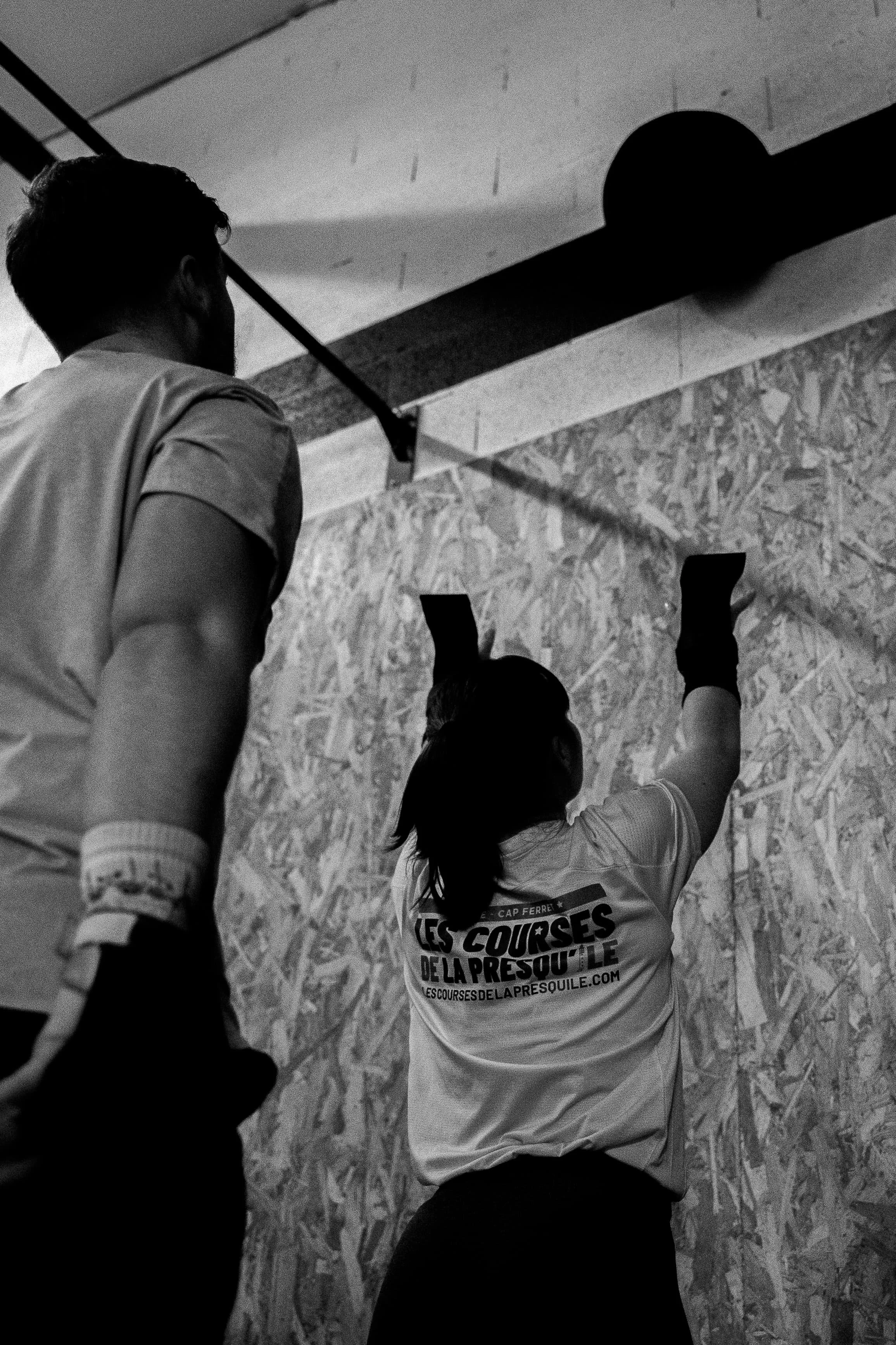 Une femme en t-shirt avec le texte 'Les courses de la Presqu'île' levée la main, vue de dos, dans une salle avec un mur en panneau de particules. À côté d'elle, un homme en chemise se tient debout. La scène est en noir et blanc.