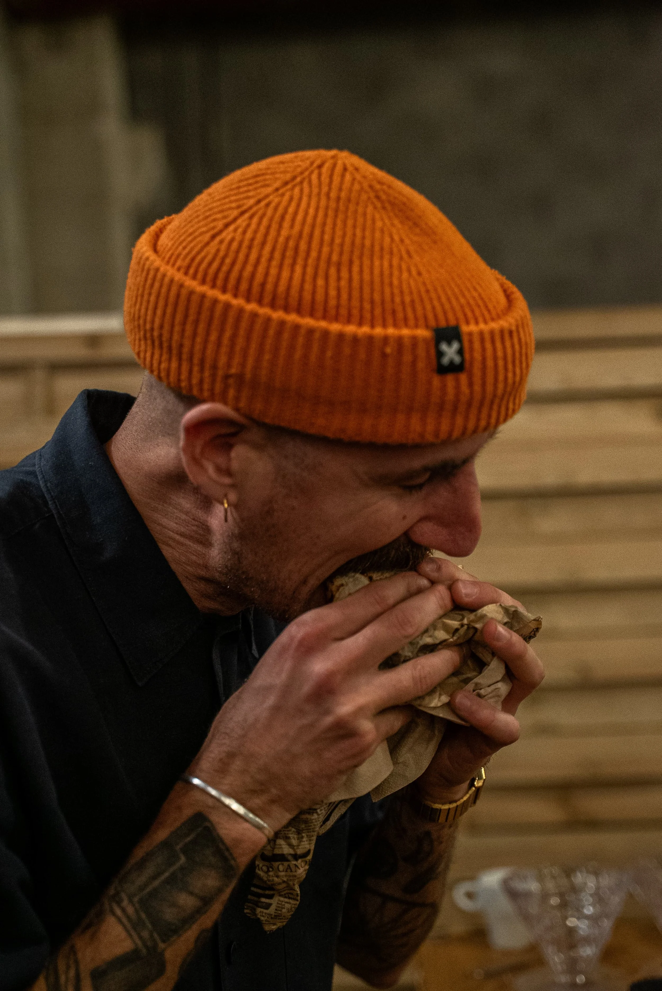 Un homme mange un sandwich dans un restaurant. Il porte un bonnet orange en tricot, un bracelet en or, une boucle d'oreille en or, et a des tatouages sur le bras.