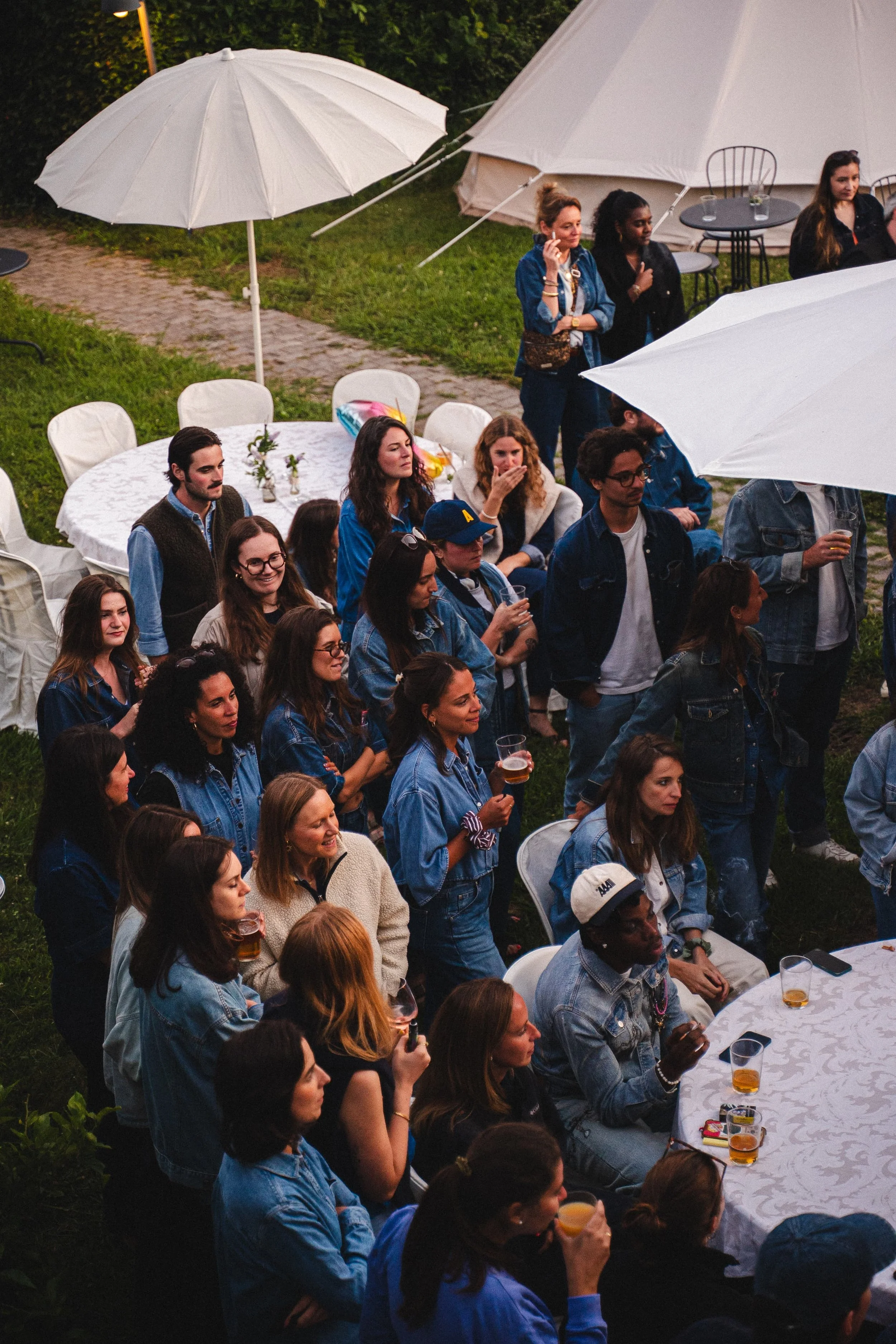 Un groupe de personnes assises autour d'une table blanche à l'extérieur lors d'une soirée ou d'un événement, avec des parasols blancs et un terrain verdoyant en arrière-plan.