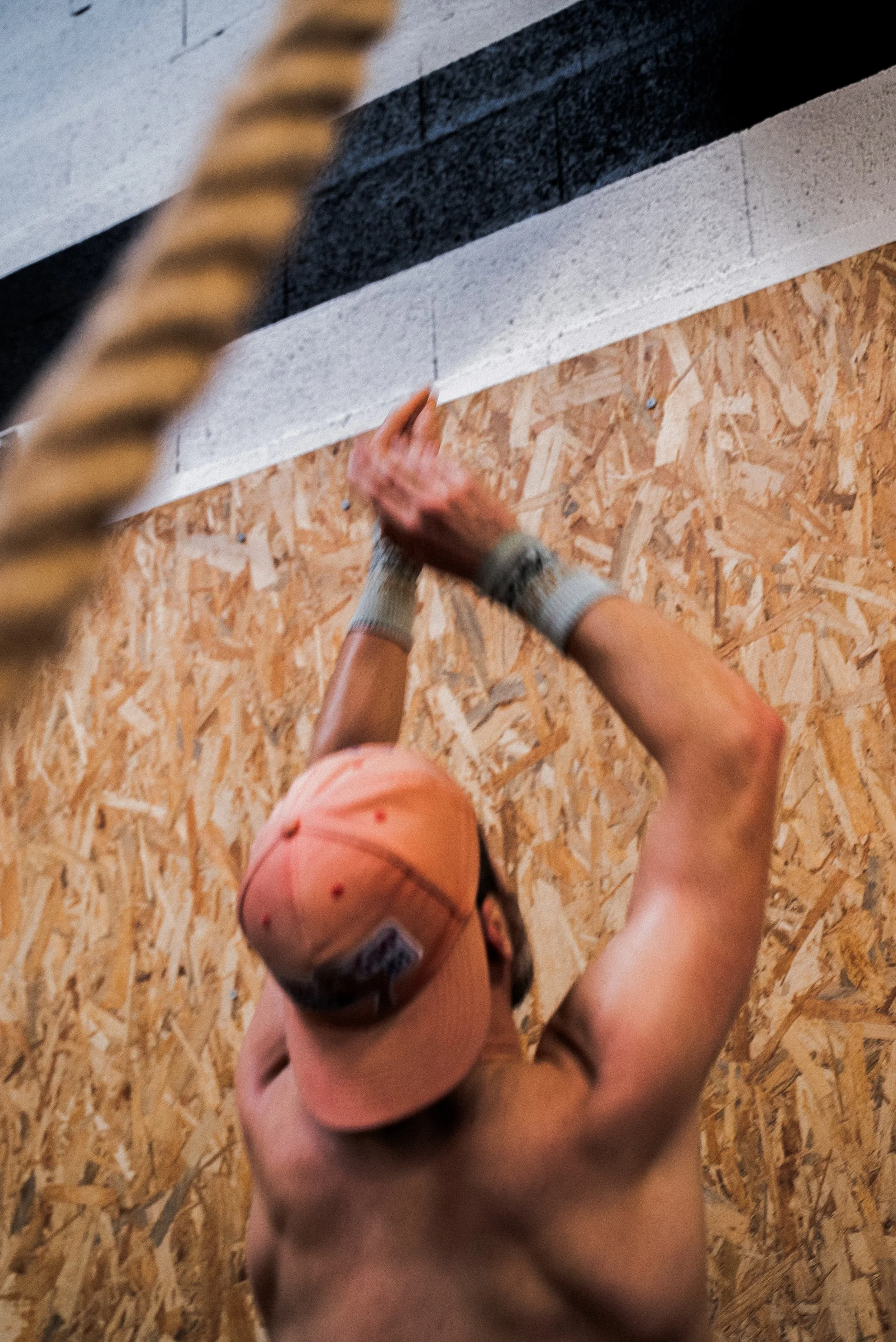 Un enfant portant une casquette orange, vu de derrière, qui monte une corde lors d'une activité en intérieur avec un mur en panneau de bois.