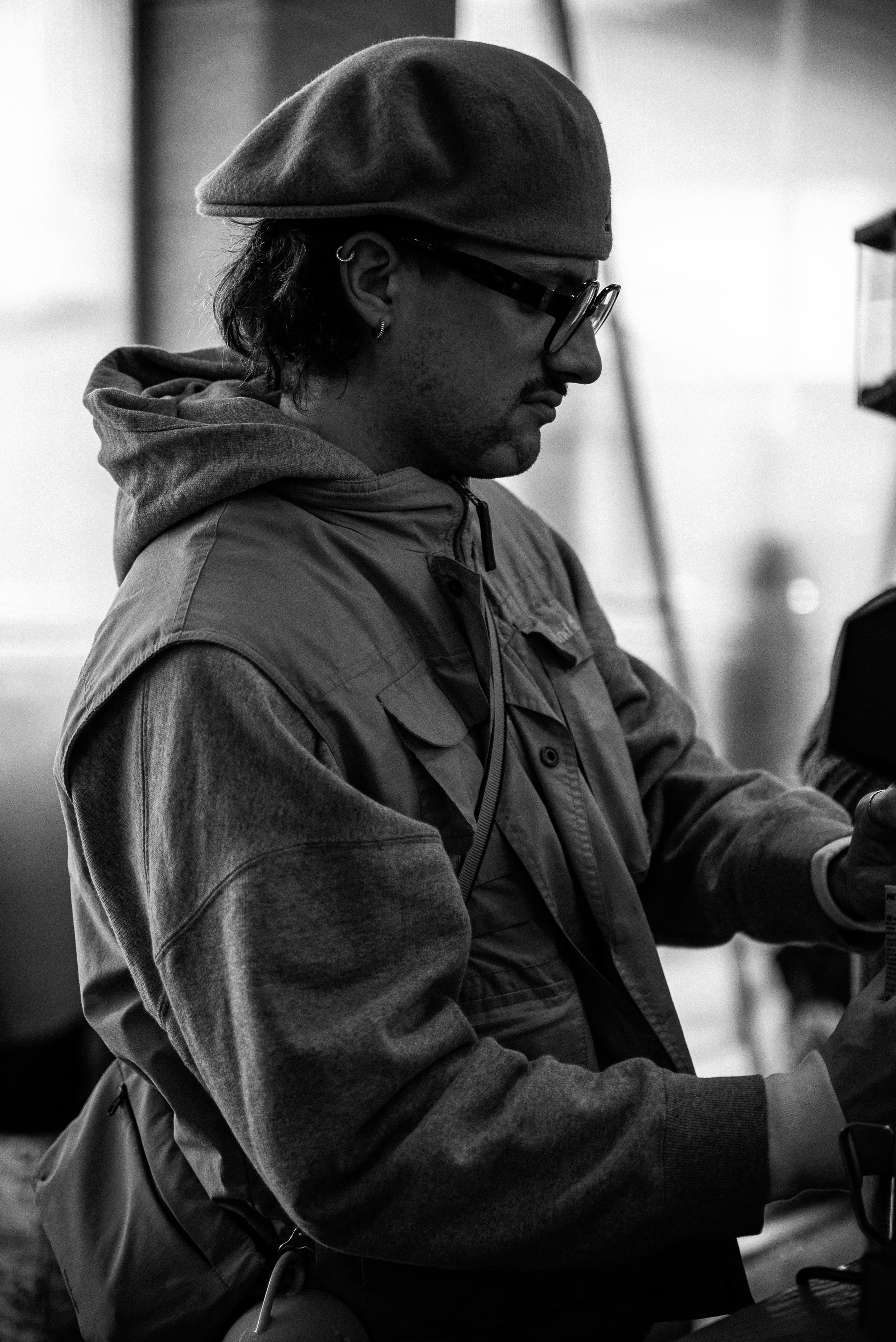 Un homme portant des lunettes, un bonnet, une veste et un sac à dos, regarde vers un appareil ou un objet qu'il tient dans ses mains, dans un environnement intérieur.