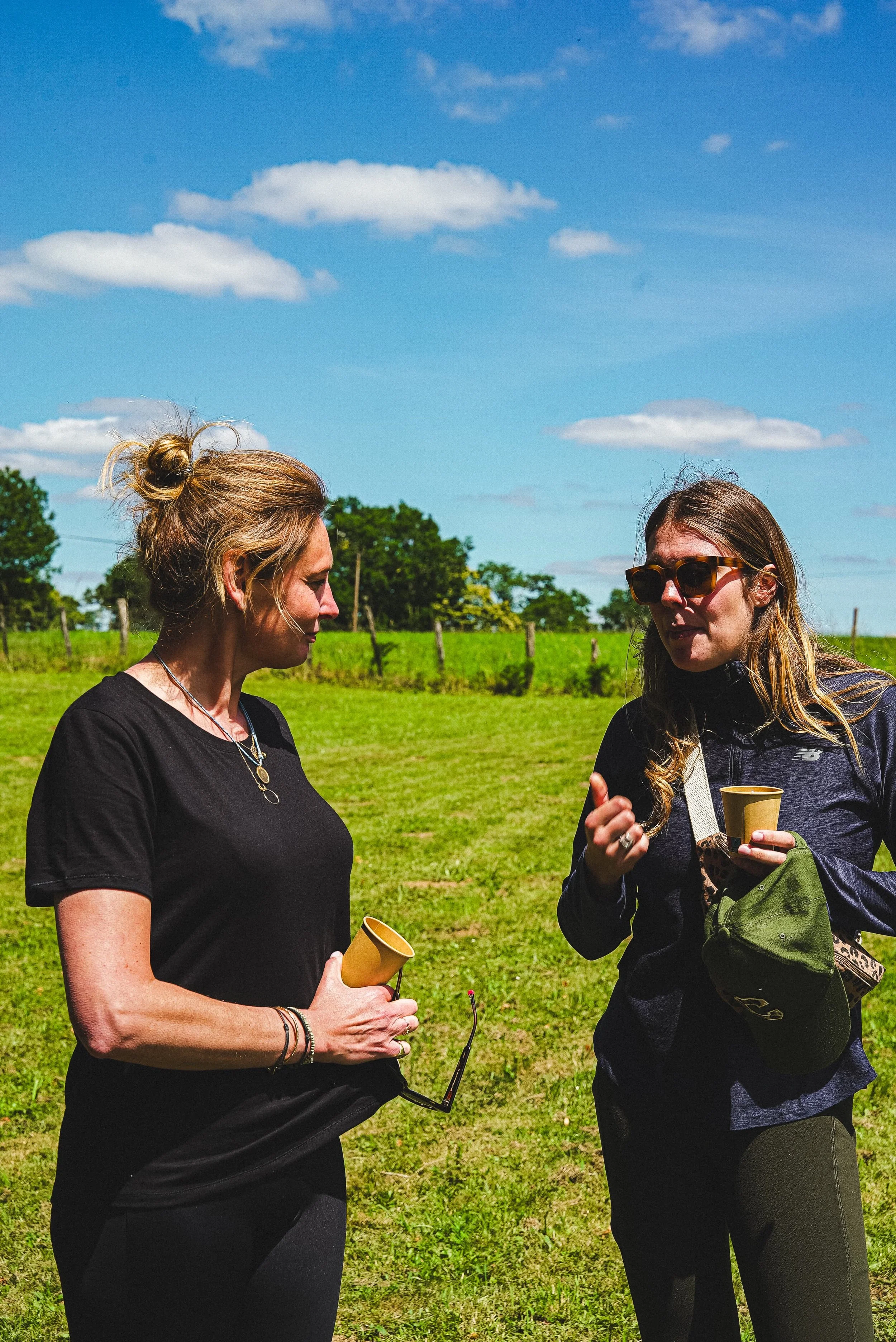 Deux femmes discutent dans un champ vert sous un ciel bleu avec quelques nuages. L'une porte des lunettes de soleil, l'autre tient une tasse en papier jaune et un petit appareil électronique, probablement un micro.