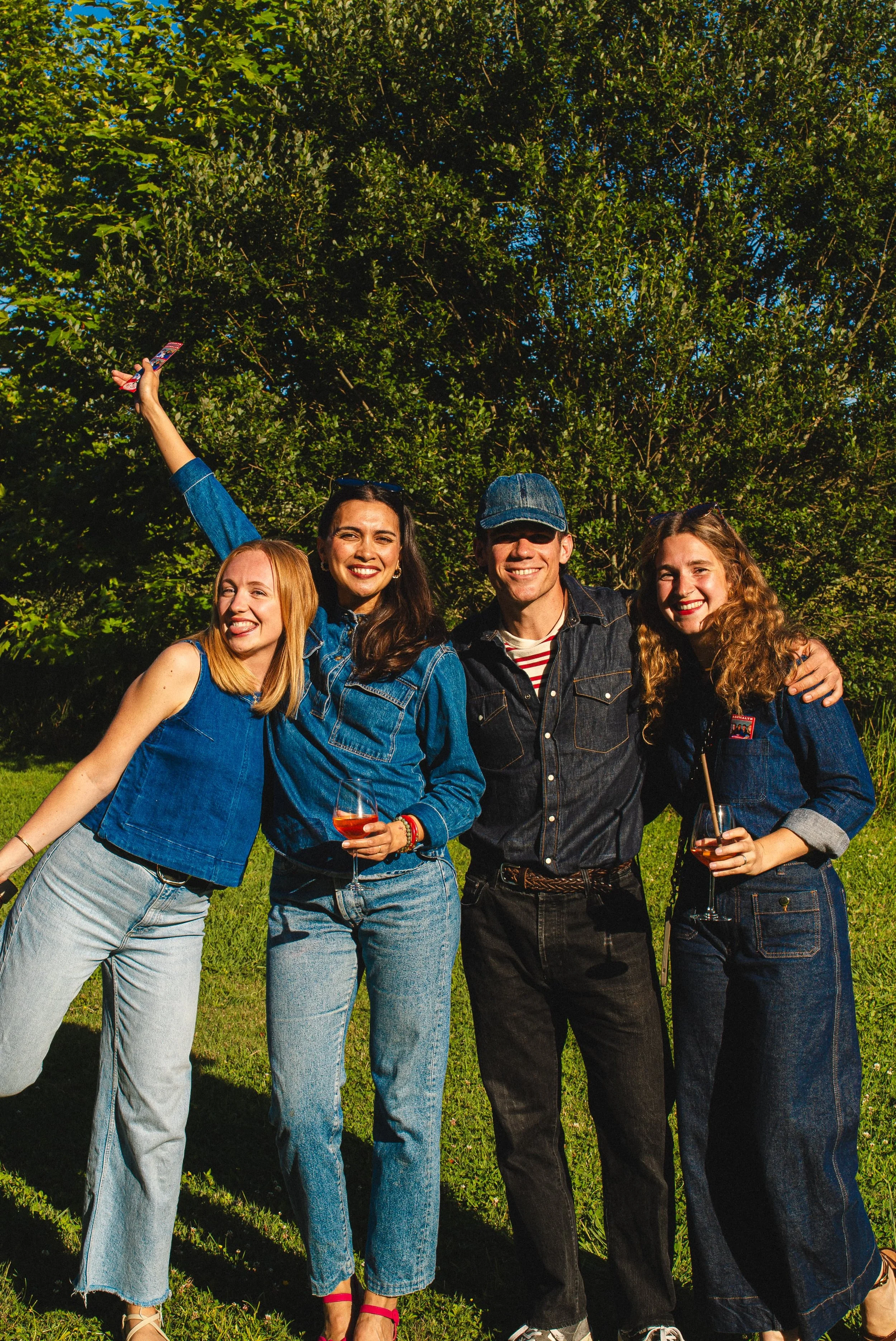 Quatre amis souriants, deux femmes et un homme, posent ensemble dans un parc ensoleillé, tenant des verres de vin rosé, entourés d'arbres verts.