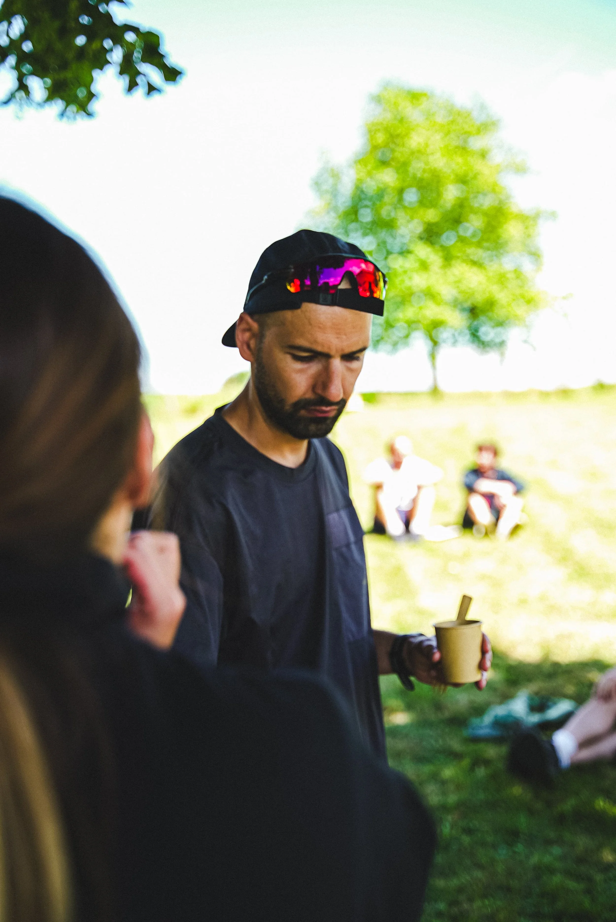 Groupe de personnes dans un parc, un homme avec une casquette et des lunettes de soleil réfléchissantes tient une tasse, d'autres personnes assises en arrière-plan, sous un arbre lumineux par une journée ensoleillée.