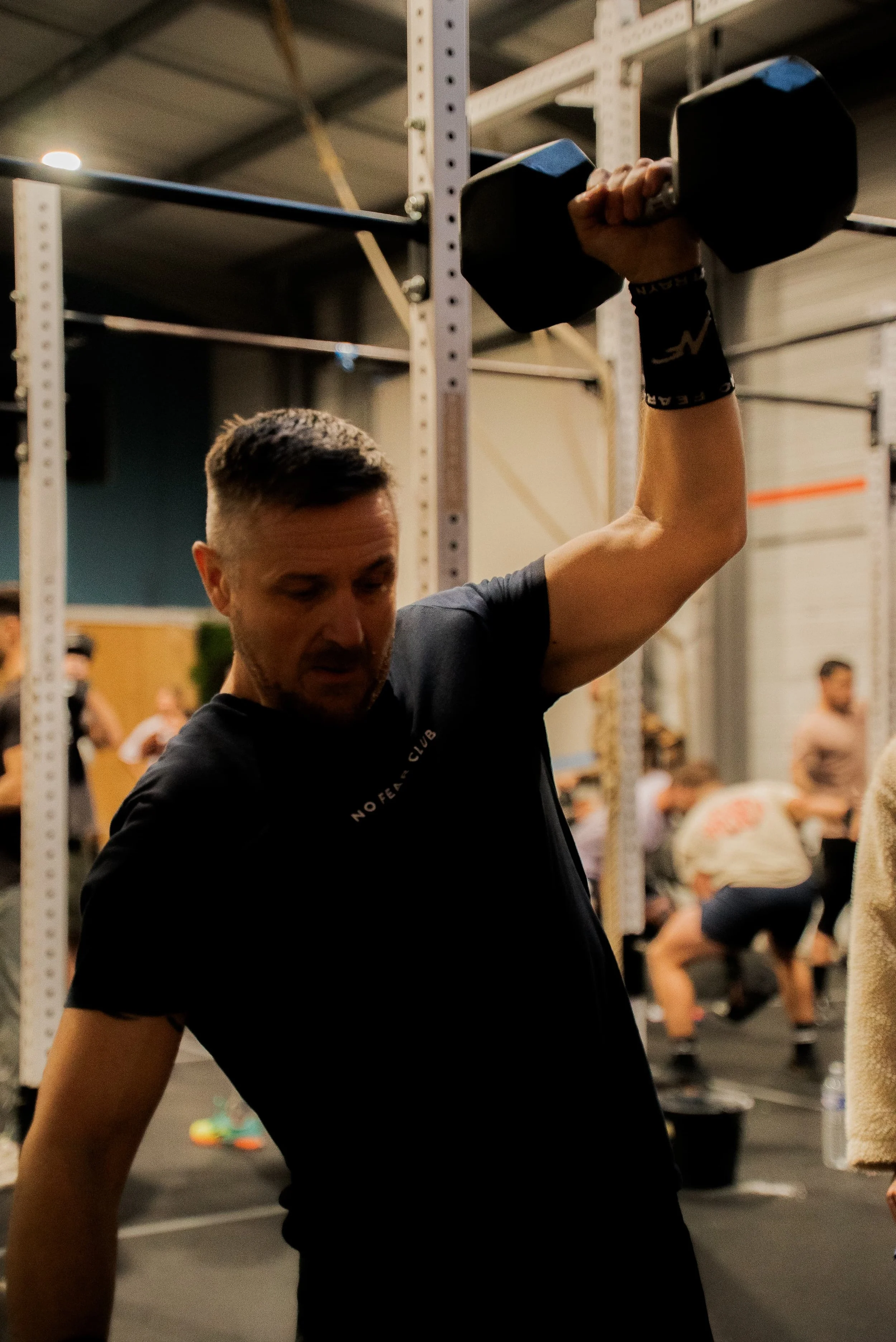 Un homme en entraînement dans une salle de sport, portant un t-shirt noir et un bracelet, soulevant un haltère noire. En arrière-plan, plusieurs personnes s'entraînent avec des équipements de musculation.