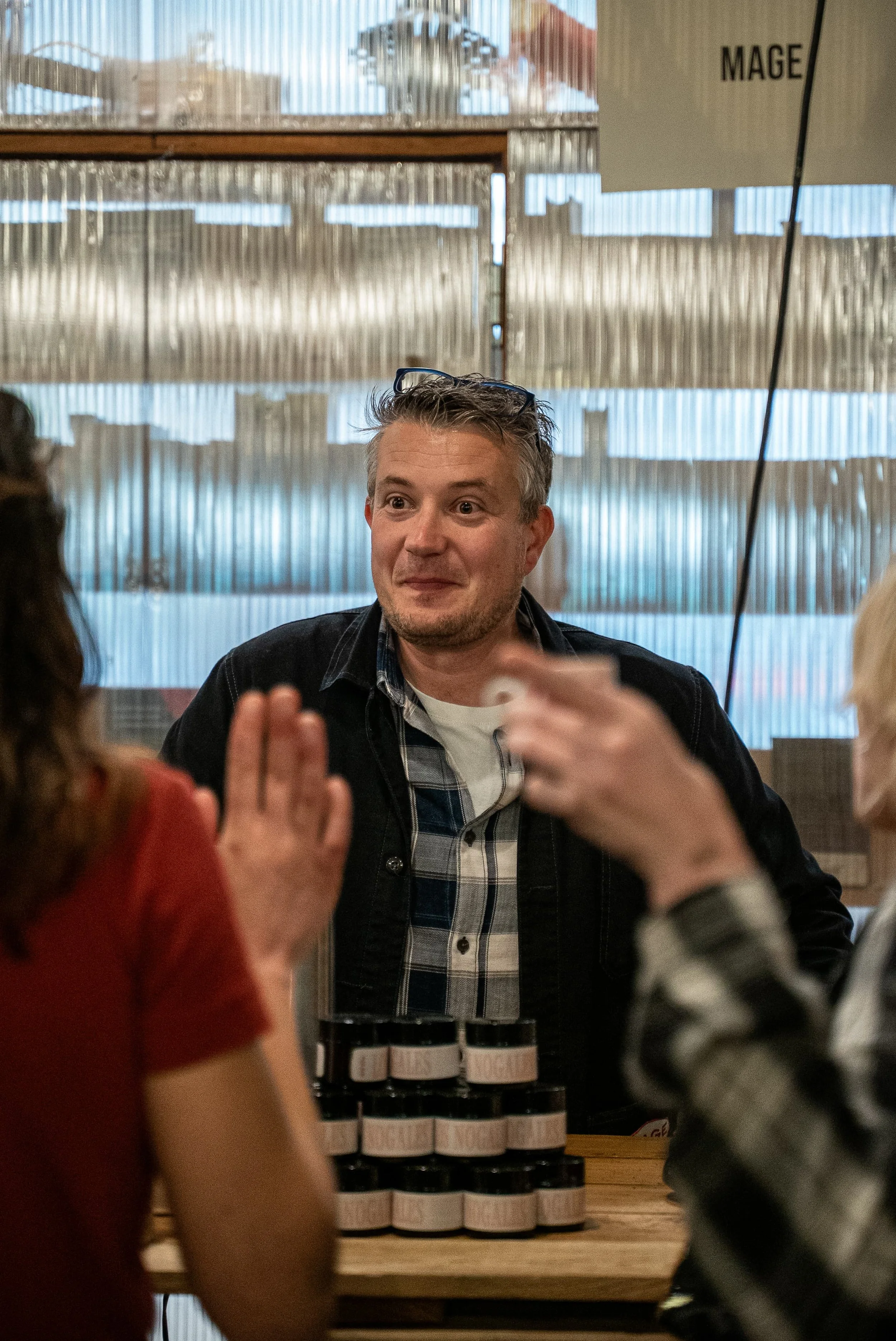 Un homme parle à un groupe de personnes dans un environnement intérieur, avec des étagères en bois et une signalisation 'MAGE' en arrière-plan.