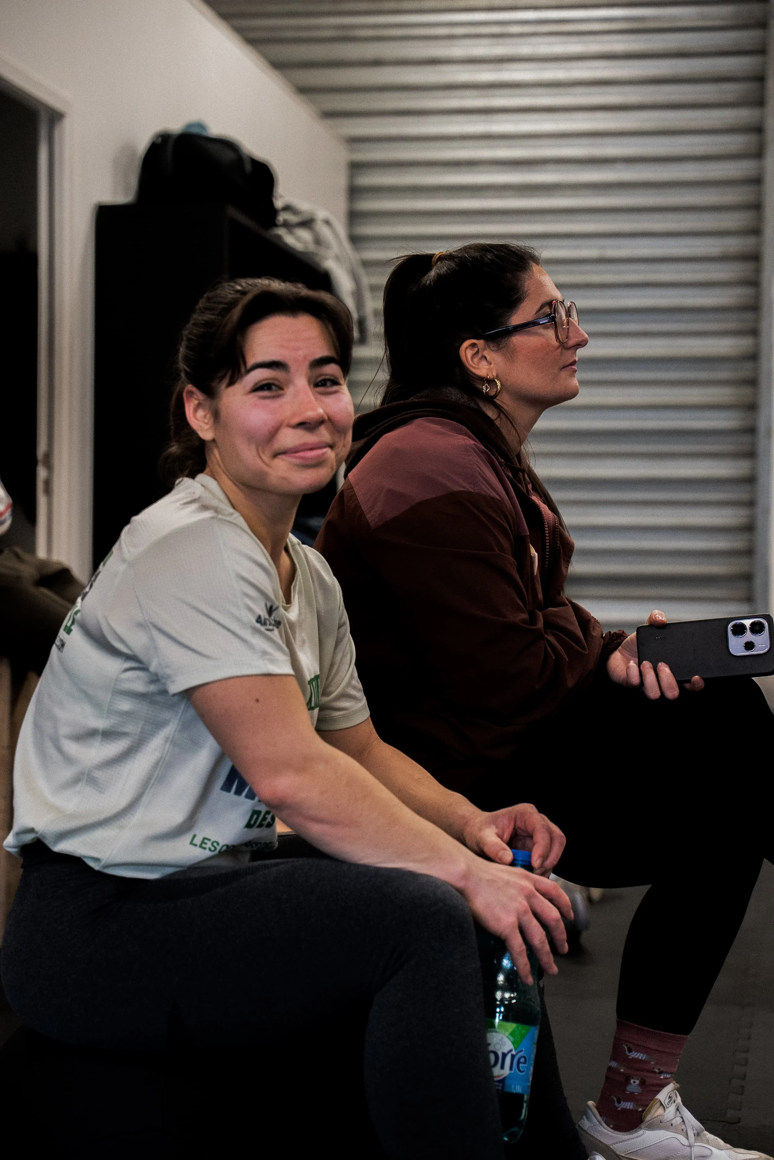 Deux femmes assises côte à côte dans une pièce, l'une souriante, l'autre regardant de côté, avec un fond de porte de garage et de casiers.