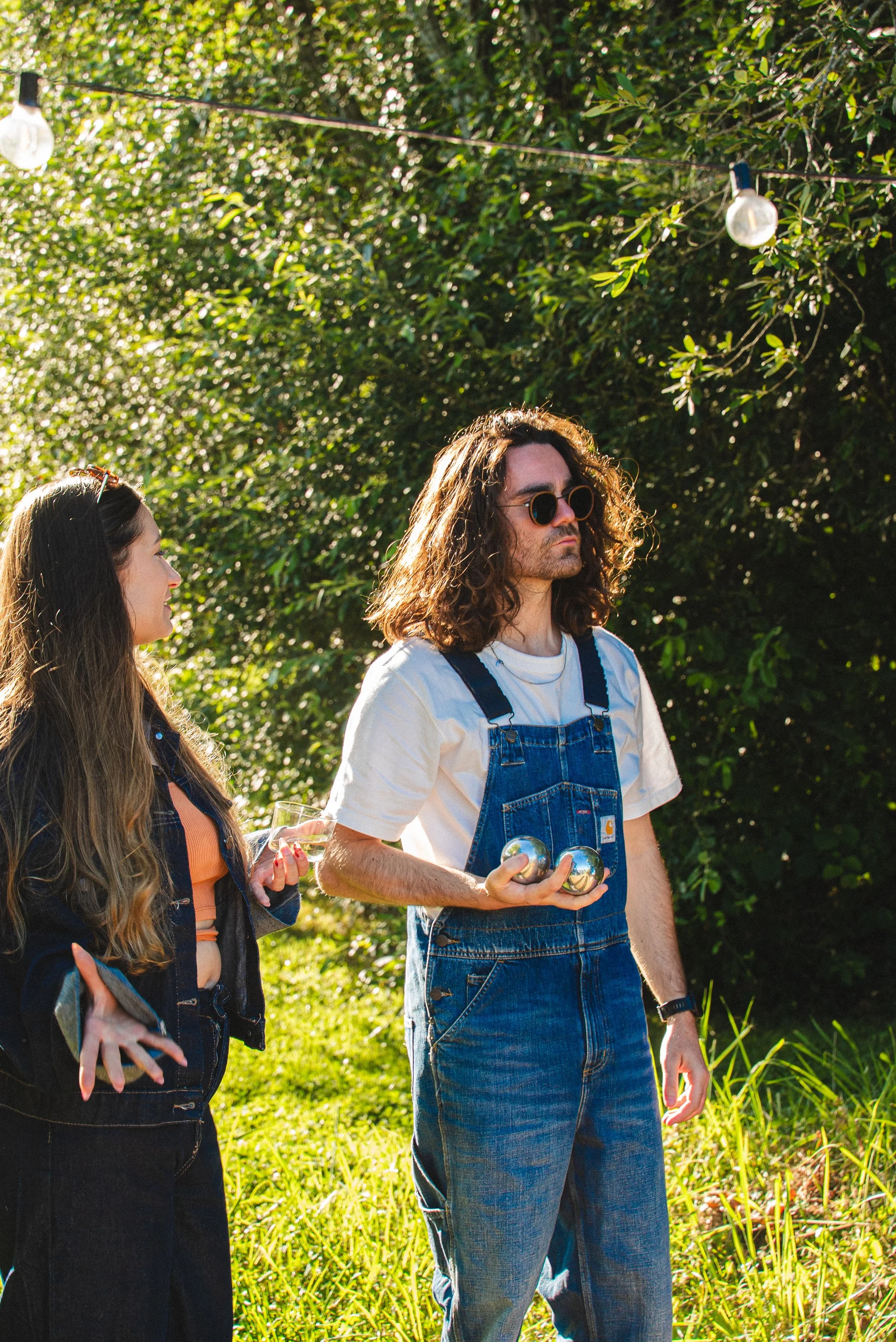 Deux personnes debout dans un jardin ensoleillé, l'une avec des lunettes de soleil et des jeans, tenant des balles métalliques, l'autre parlant, portant une veste en jean et un t-shirt orange, avec des lumières décoratives suspendues au-dessus.