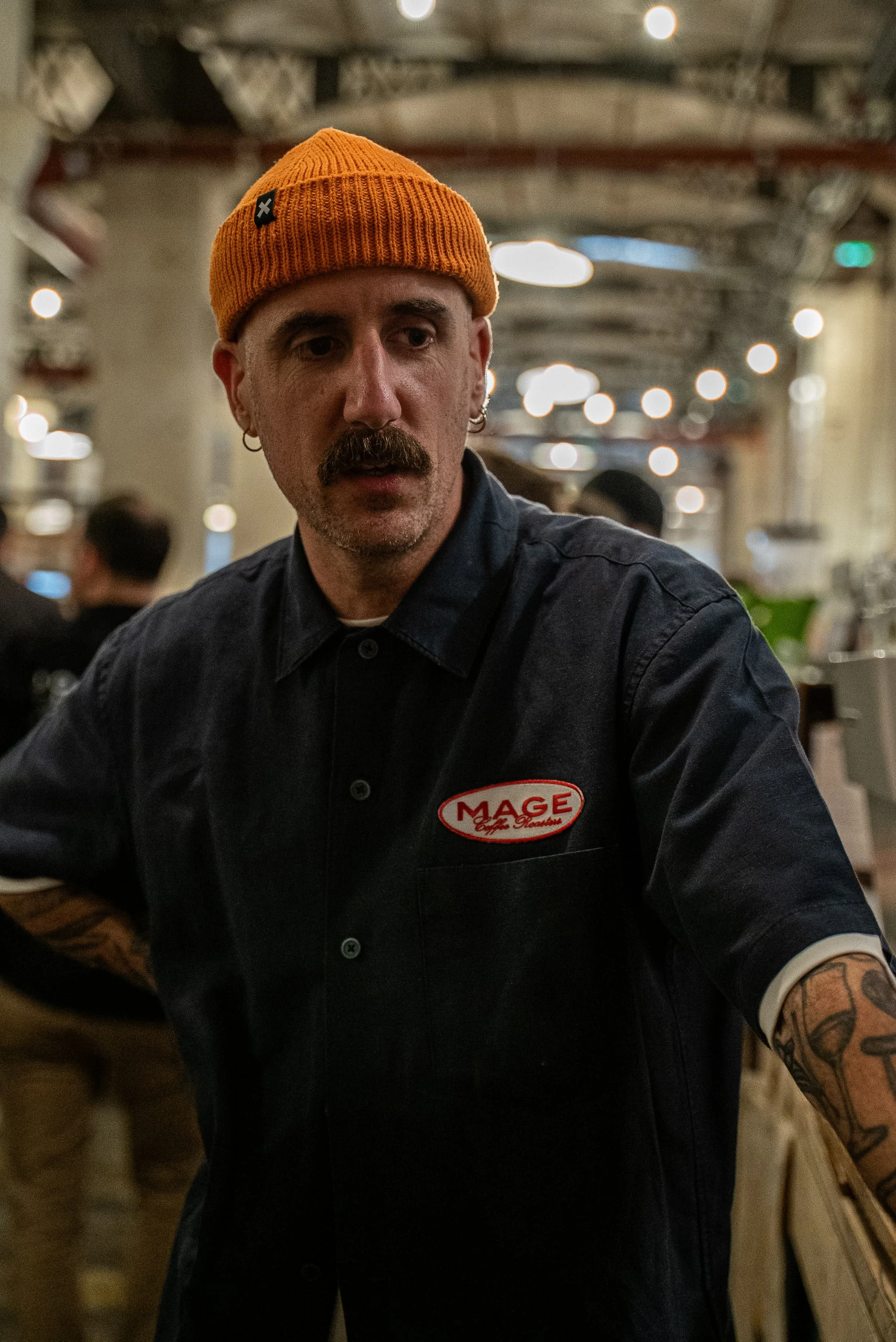 Un homme avec une barbe, moustache, et cheveux courts portant un bonnet orange et une chemise noire avec un badge "MAGE" observe quelque chose dans un lieu intérieur éclairé avec des personnes en arrière-plan.