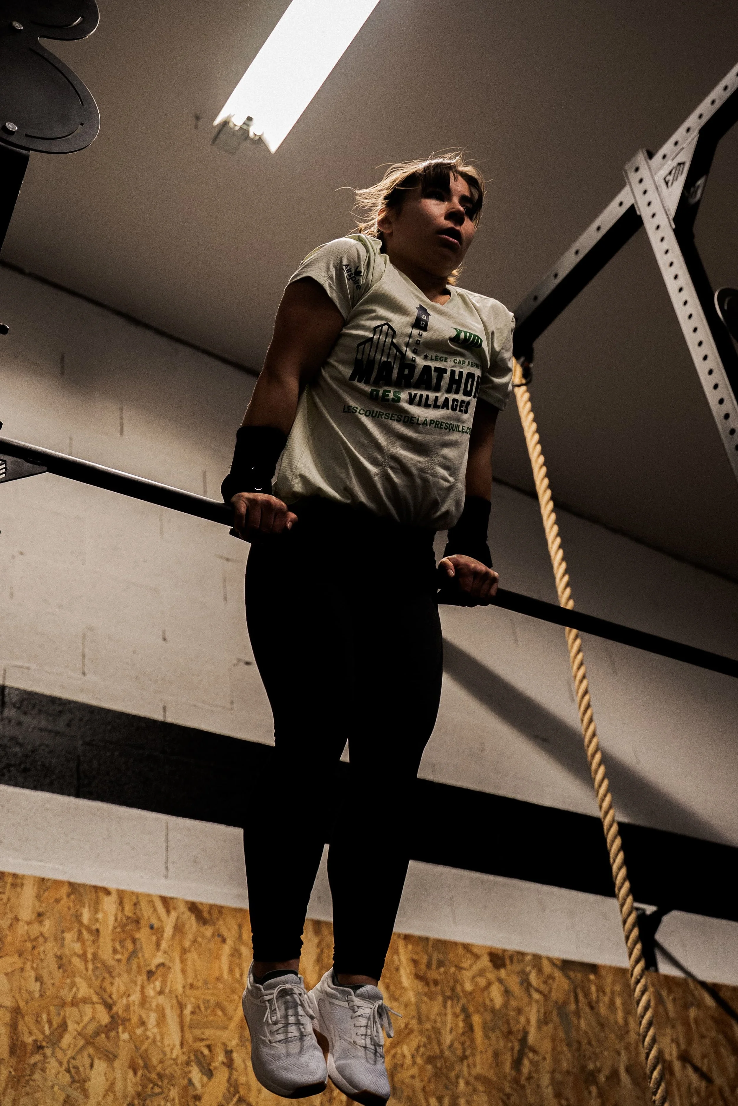 Une femme en tenue de sport suspendue à une barre d'entraînement, portant des baskets blanches, dans une salle de sport avec un mur en bois et une corde d'entraînement à côté.
